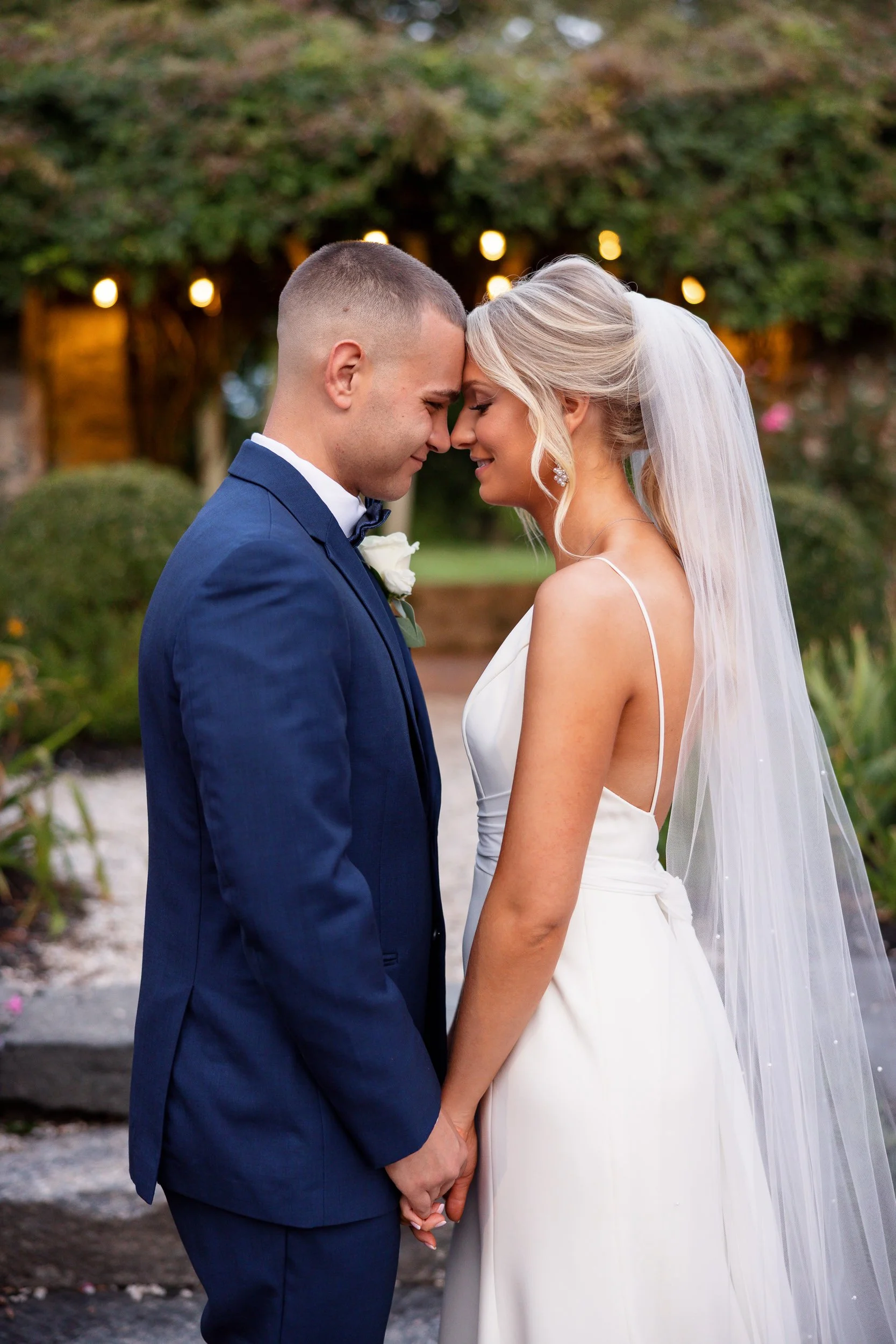 A bride and groom holding hands with their foreheads together, garden lights soft behind them.
