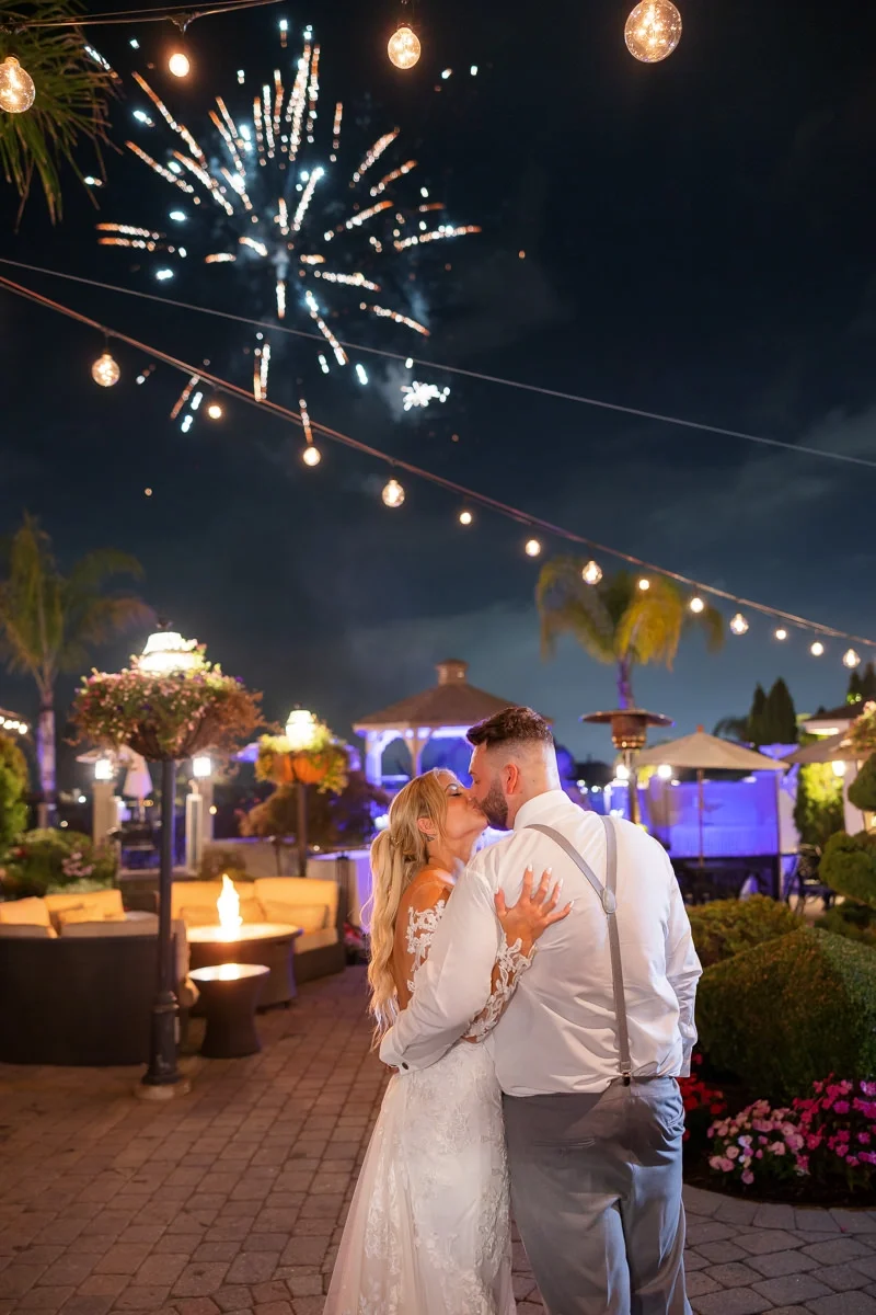 A couple is kissing at night during a celebration with fireworks in the sky. String lights are hanging overhead, and there are outdoor furniture and lush plants around them.