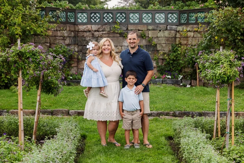 Family of four standing on a garden pathway, smiling, with flowering bushes and a stone wall in the background. The mother is holding a young girl, and the father has a young boy beside him.