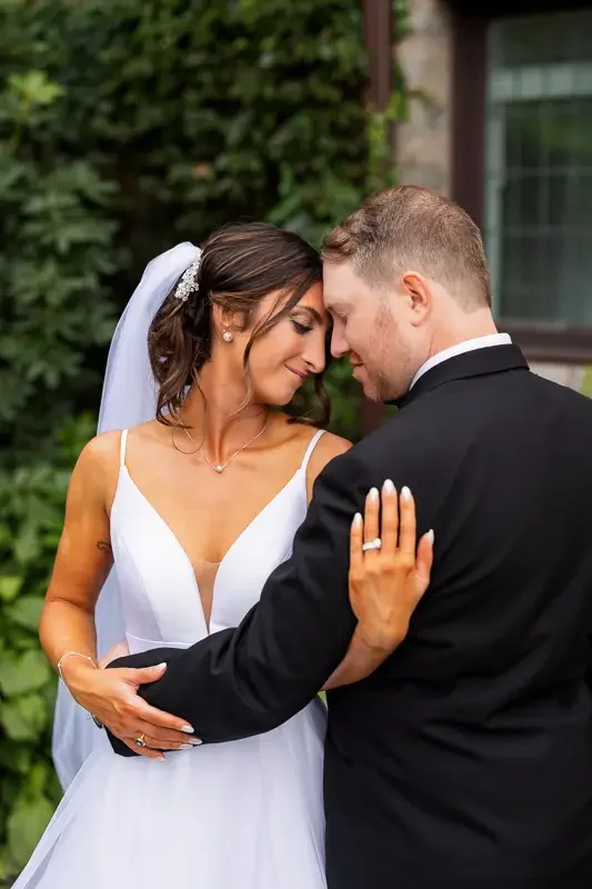A bride and groom sharing an intimate moment, with their foreheads touching and eyes closed, outdoors with green foliage and a house in the background.