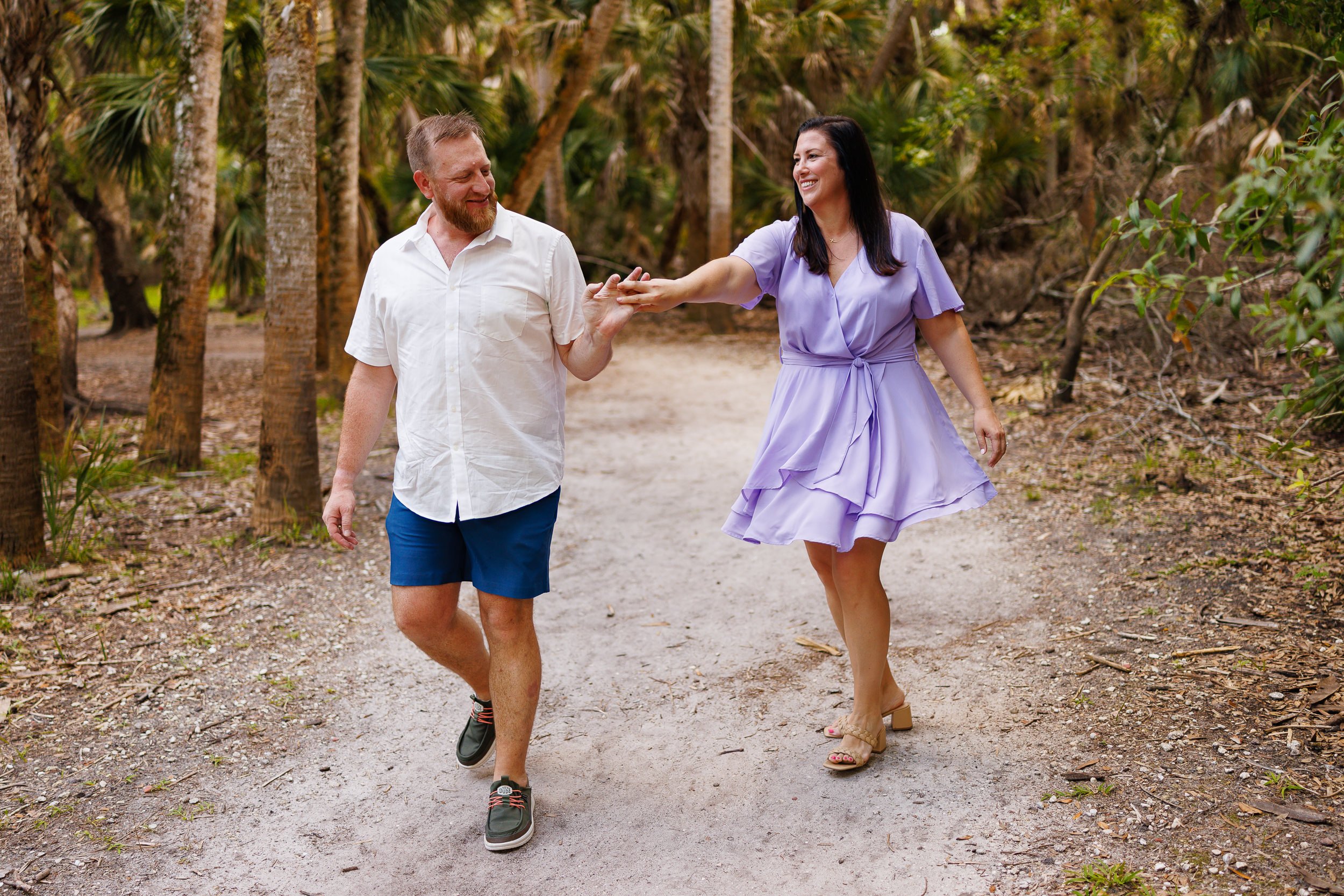 Erin reaching for Will's hand as they walk the palm-lined trail toward the Myakka canopy walkway, both laughing.