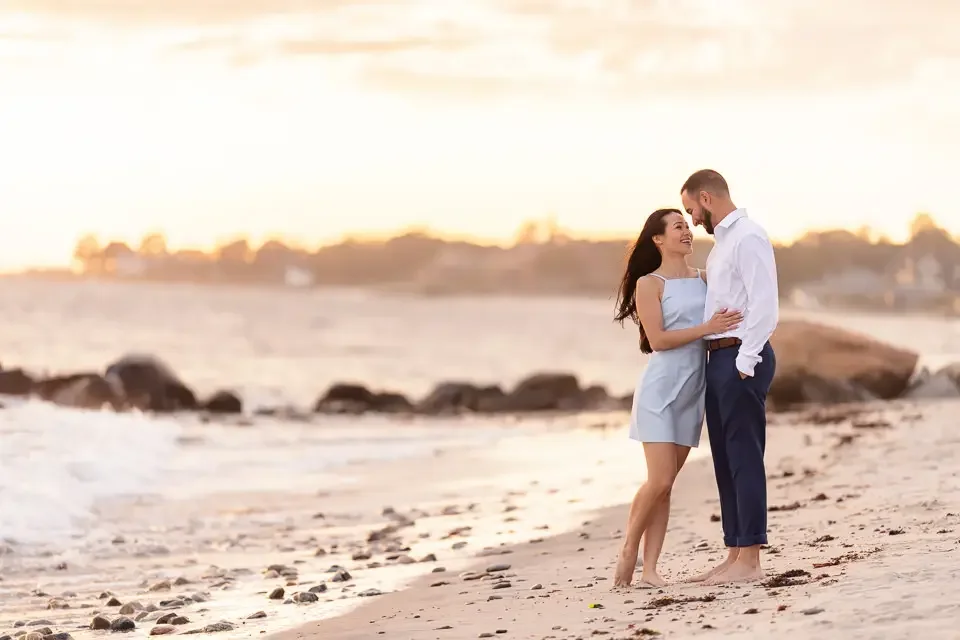 A couple standing barefoot along the shoreline during golden hour, wrapped in soft light and quiet intimacy during their beach engagement session.
