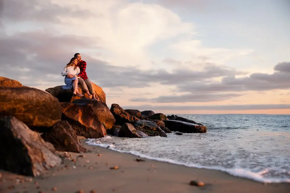 A romantic engagement session at the beach as the couple sits together on coastal rocks, surrounded by soft ocean waves and a pastel sunset sky.