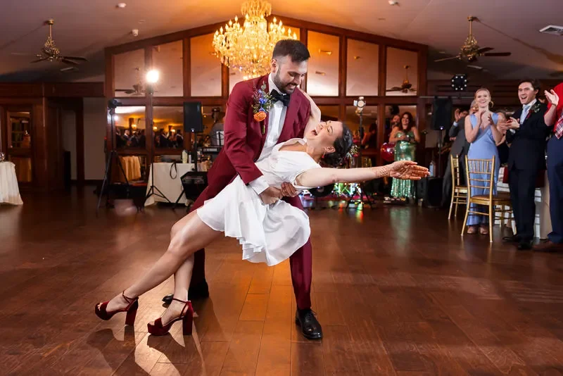 Couple dancing at a wedding reception, with the groom in a burgundy suit and the bride in a white dress, surrounded by guests clapping and celebrating in a decorated hall with chandeliers.