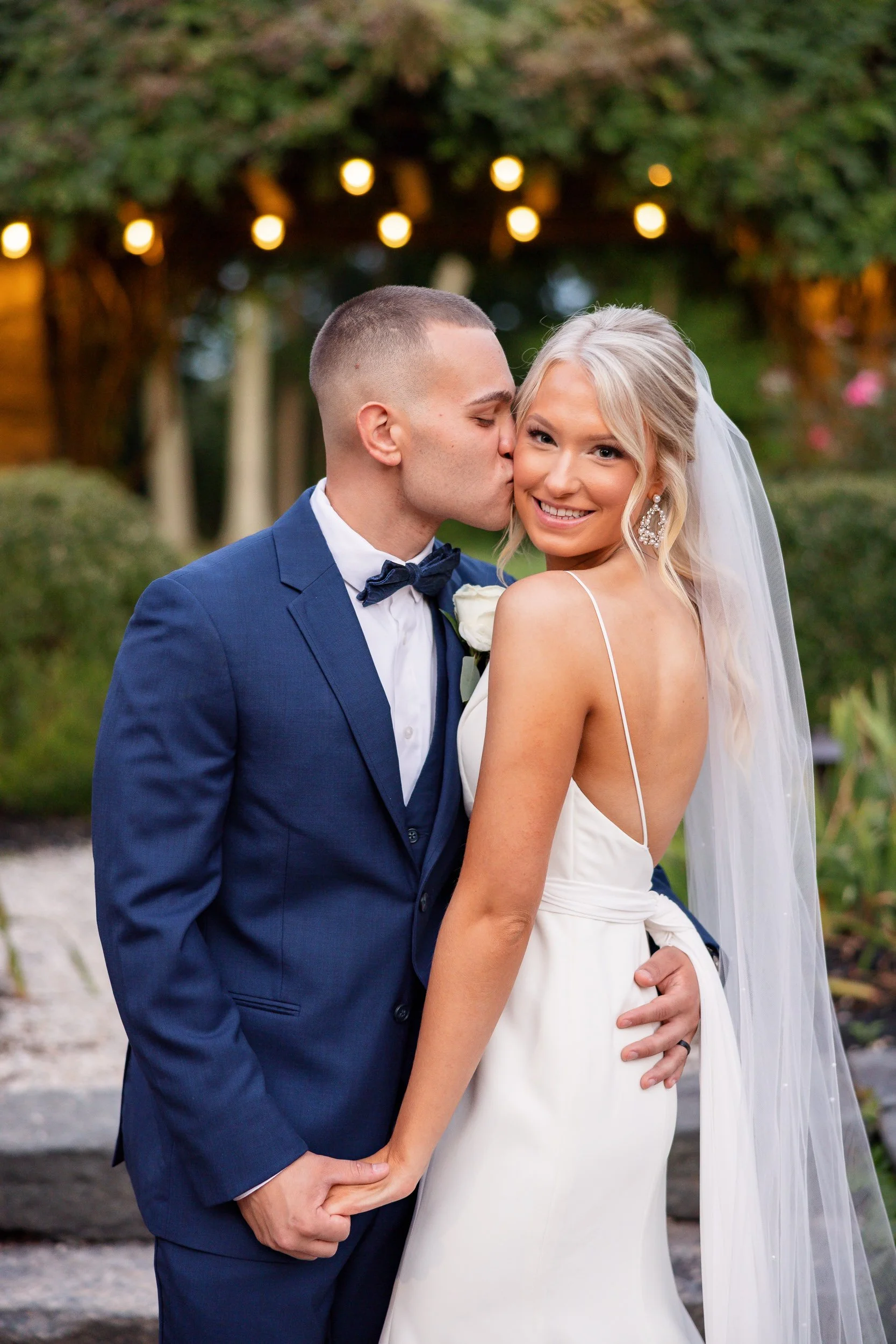 A groom in a navy suit kissing his bride's cheek in a garden, string lights strung overhead.