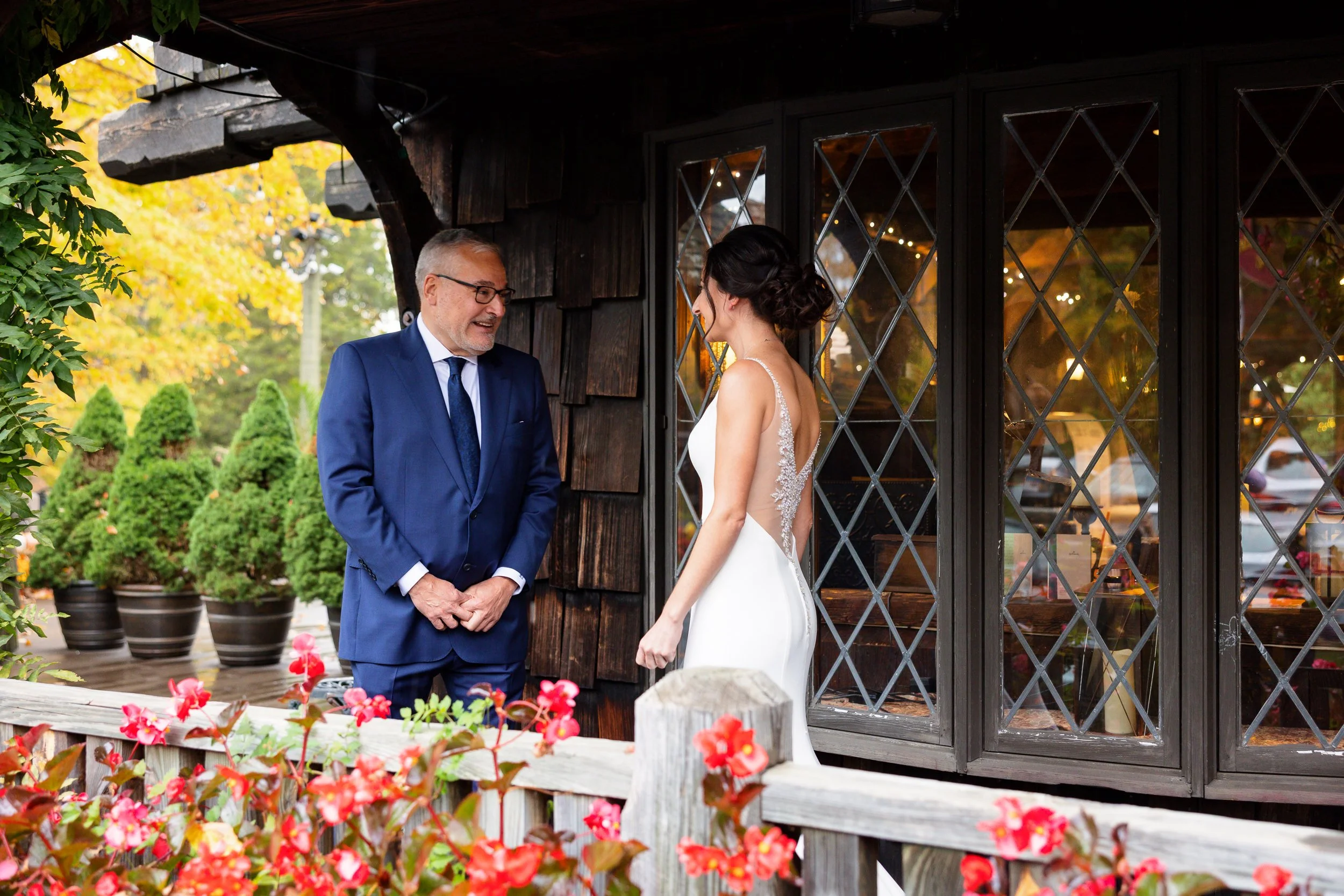 A bride's father seeing her for the first time on a covered porch, autumn leaves and red begonias around them.