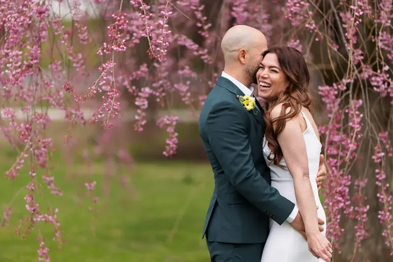 A bride and groom sharing a kiss outdoors surrounded by blooming pink flowers.