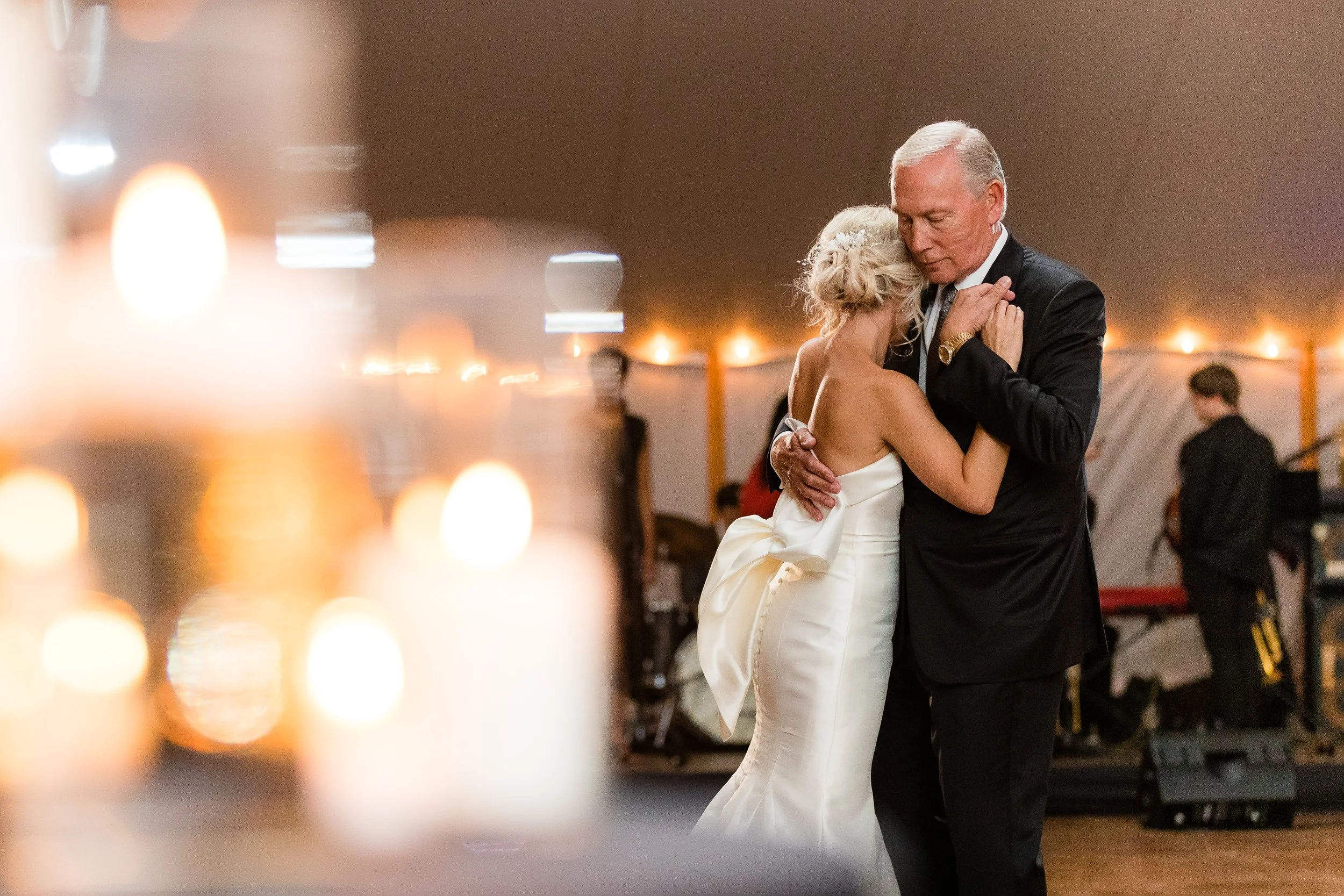 A bride dancing with her father under a tent, string lights and candles out of focus in the foreground.