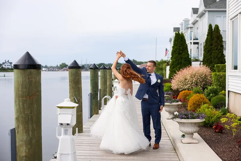 A bride and groom dancing on a dock by the water, with houses and trees in the background.