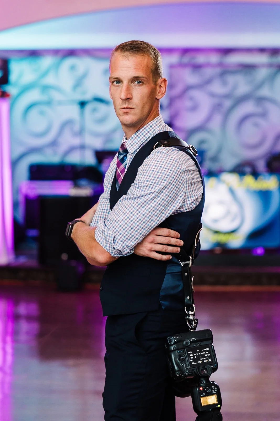 A man in business attire standing with arms crossed in a colorful, illuminated room, with a camera hanging from his shoulder.