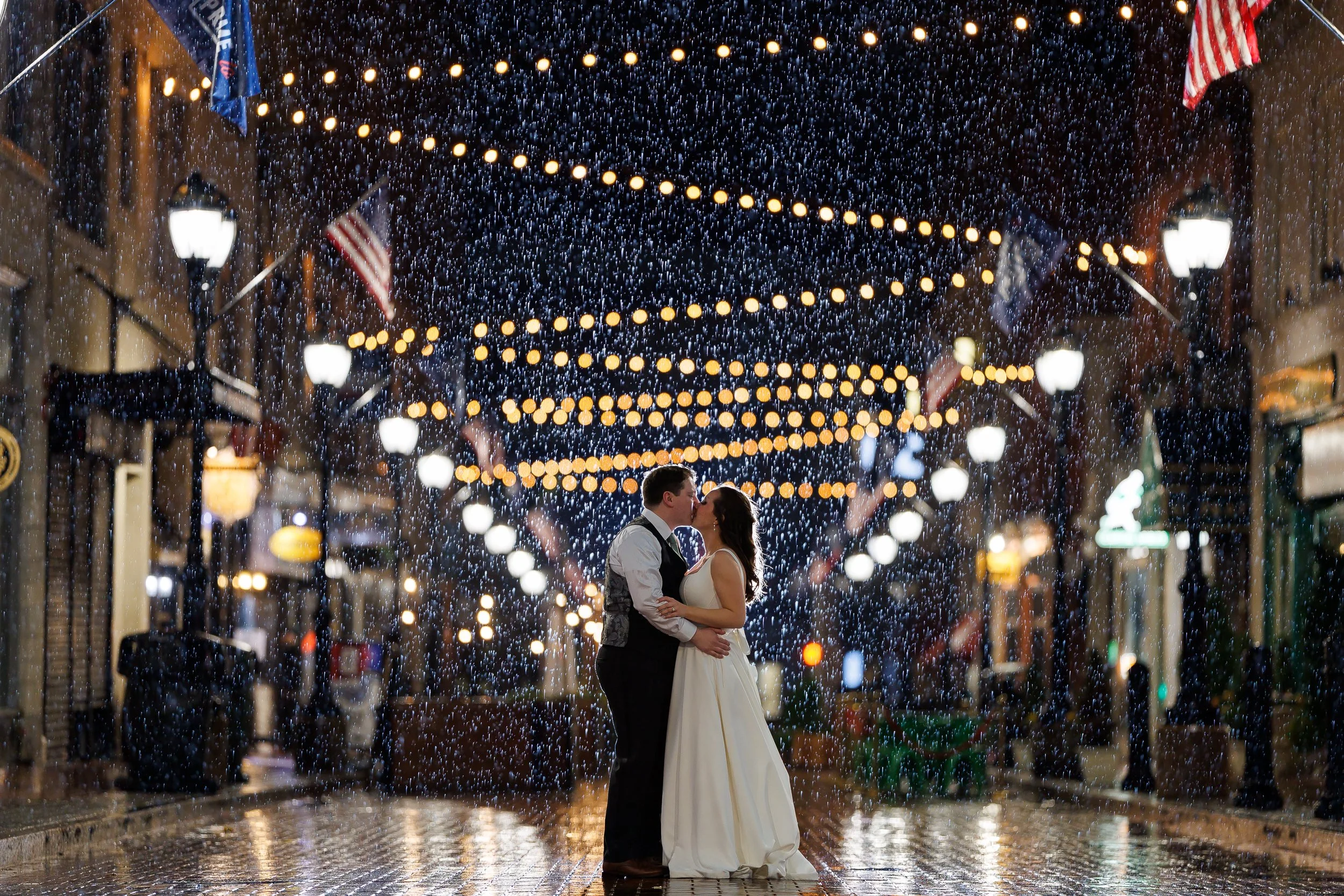 A bride and groom kissing in the rain beneath rows of string lights on a city street at night.