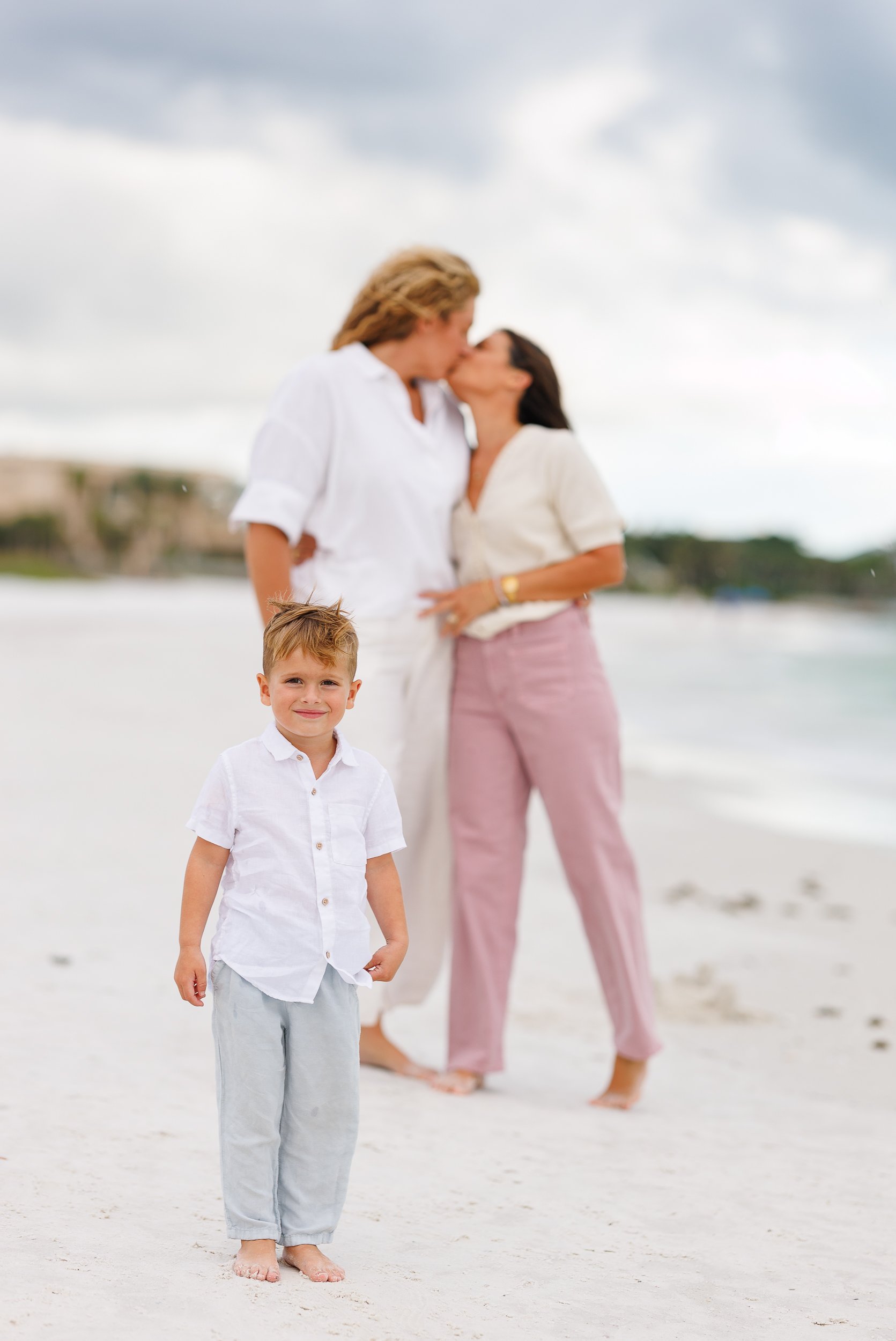A child on white sand under a clouded sky as two parents kiss behind them, Siesta Key