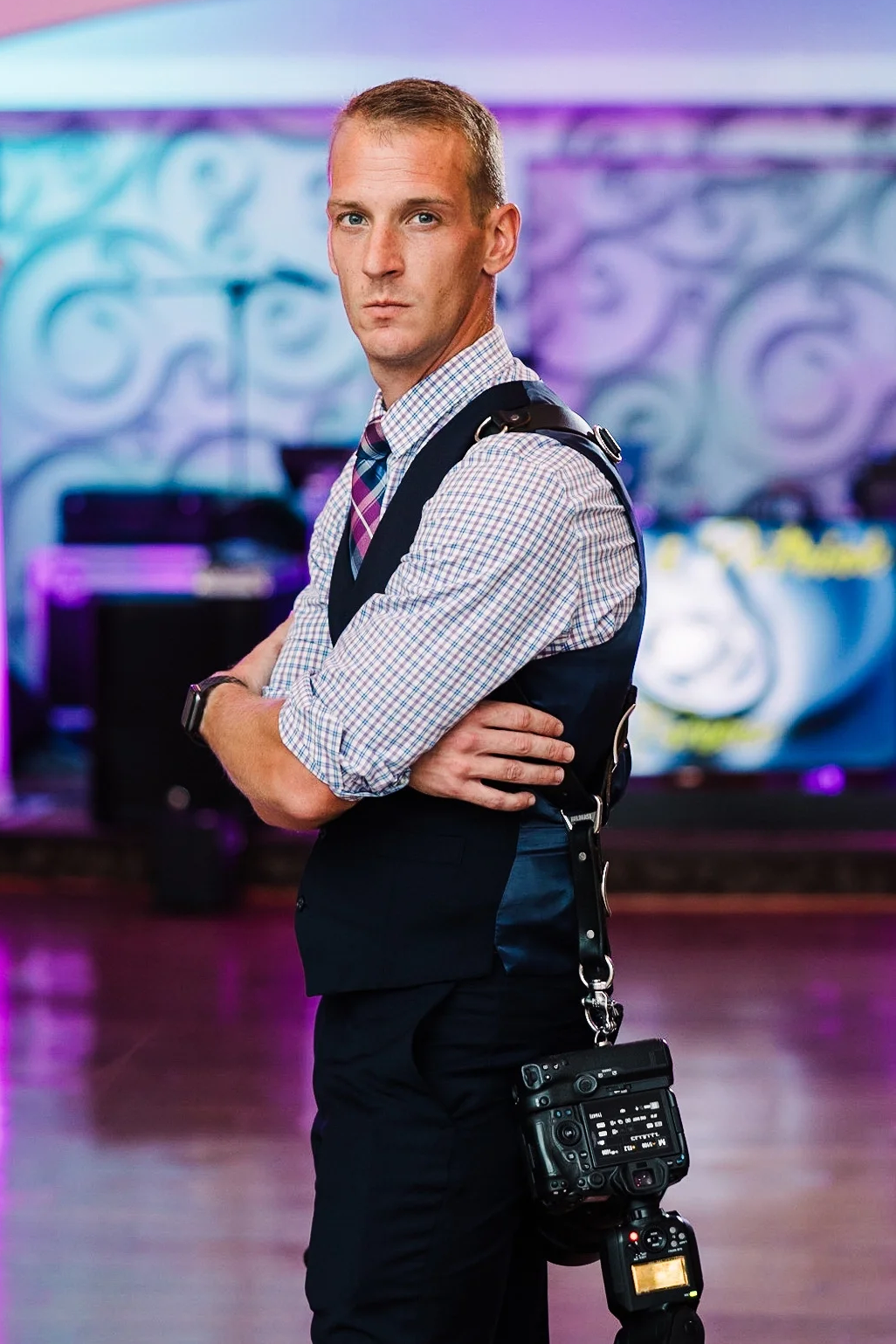 A man in business attire with a camera hanging from his neck, standing with arms crossed in a colorful indoor setting.