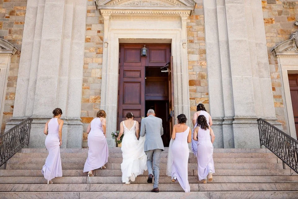 A couple walking toward a church door surrounded by attendants in lavender dresses.