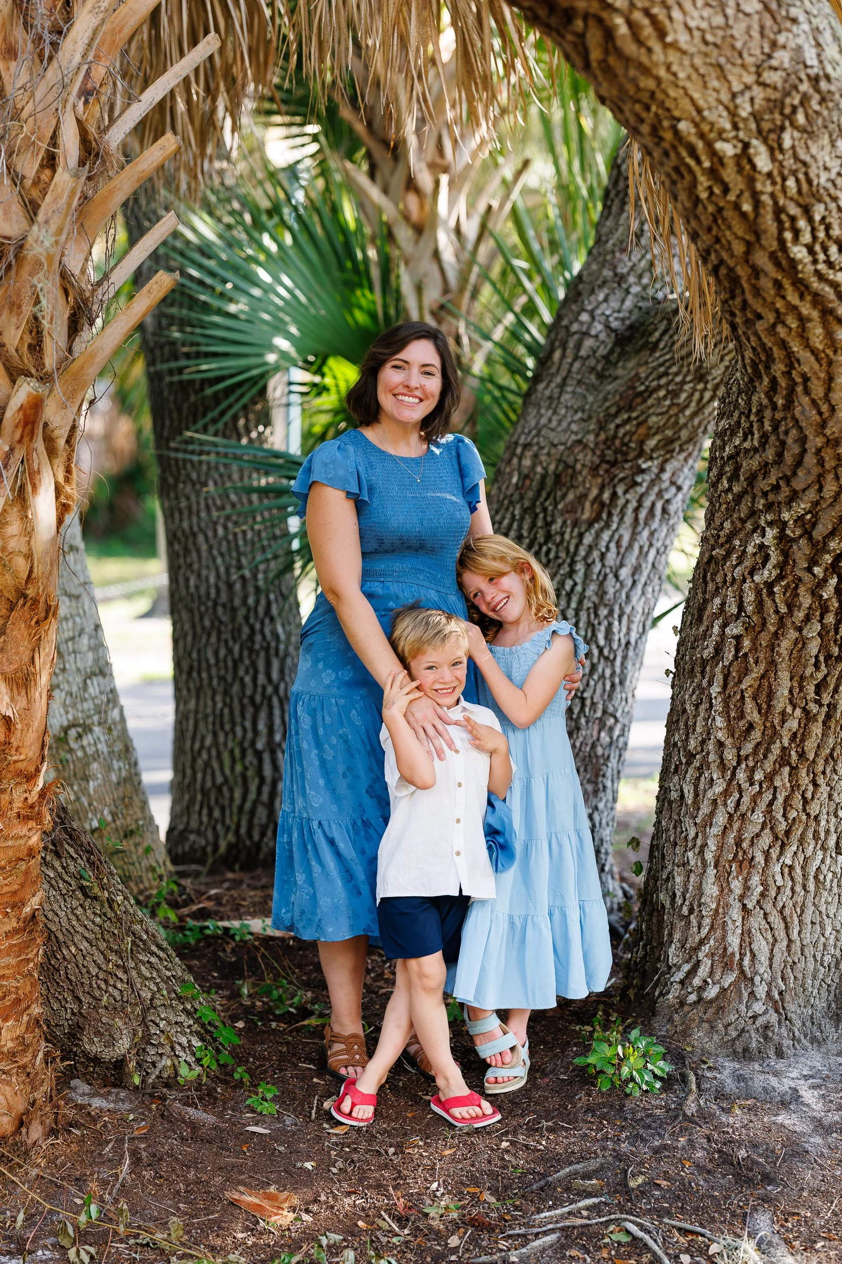 A person in a blue dress stands with two children under large trees.