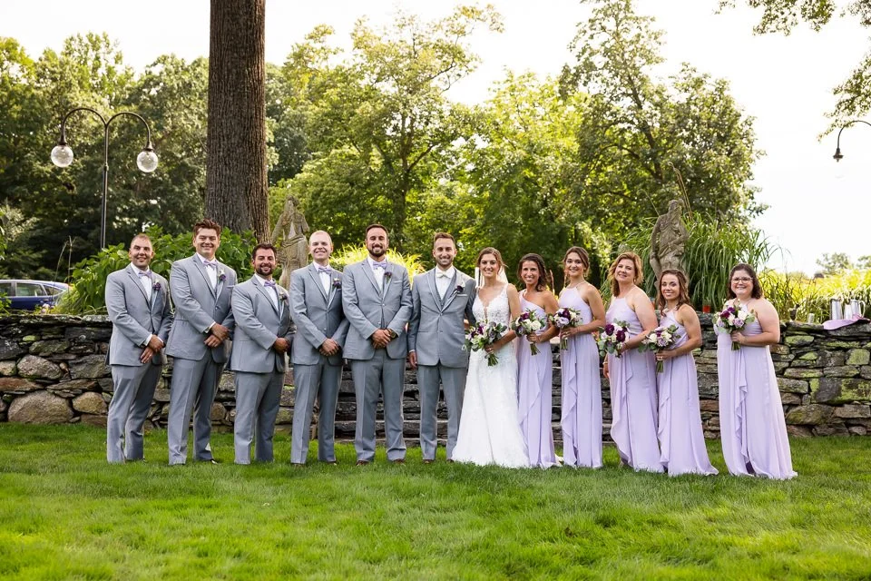 Wedding party of six people in gray suits and six in lavender dresses lined up on a lawn.