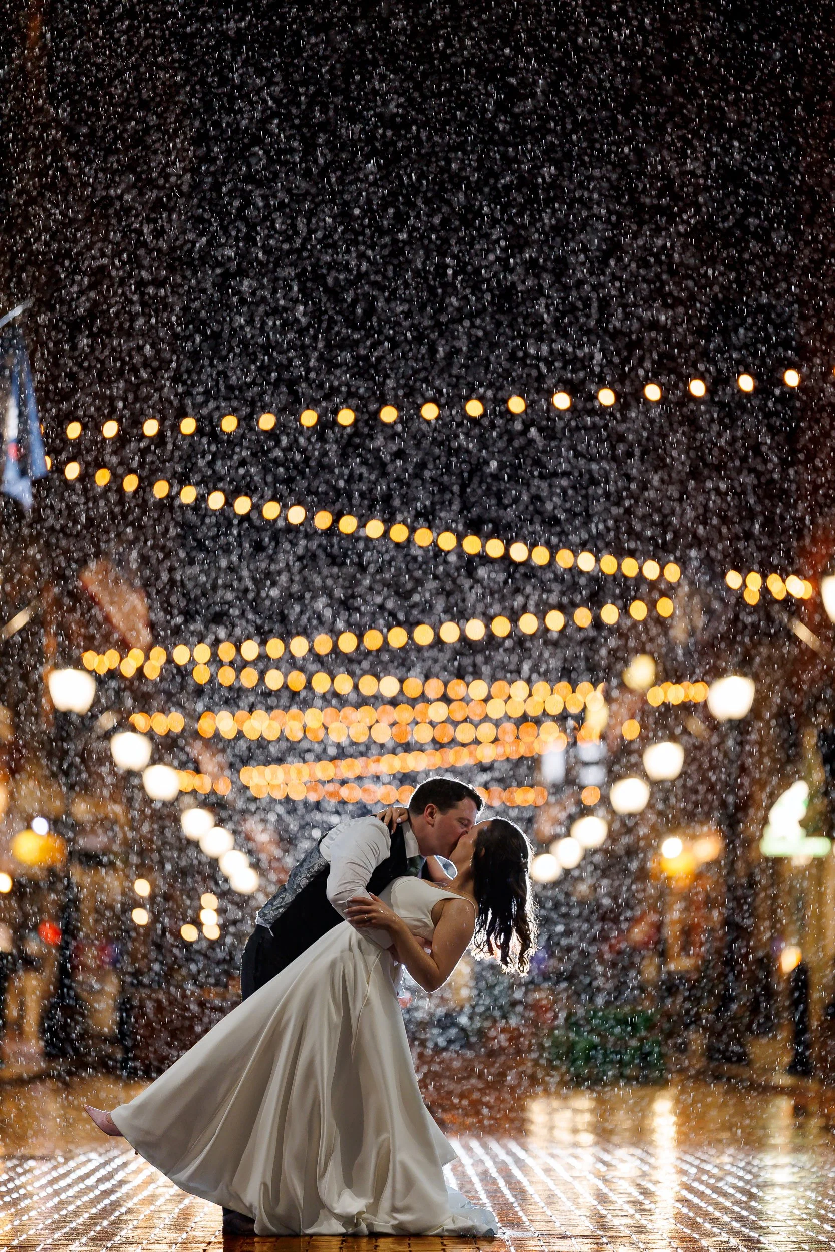 A groom dipping his bride for a kiss in heavy rain, string lights above them and the wet street reflecting the light.