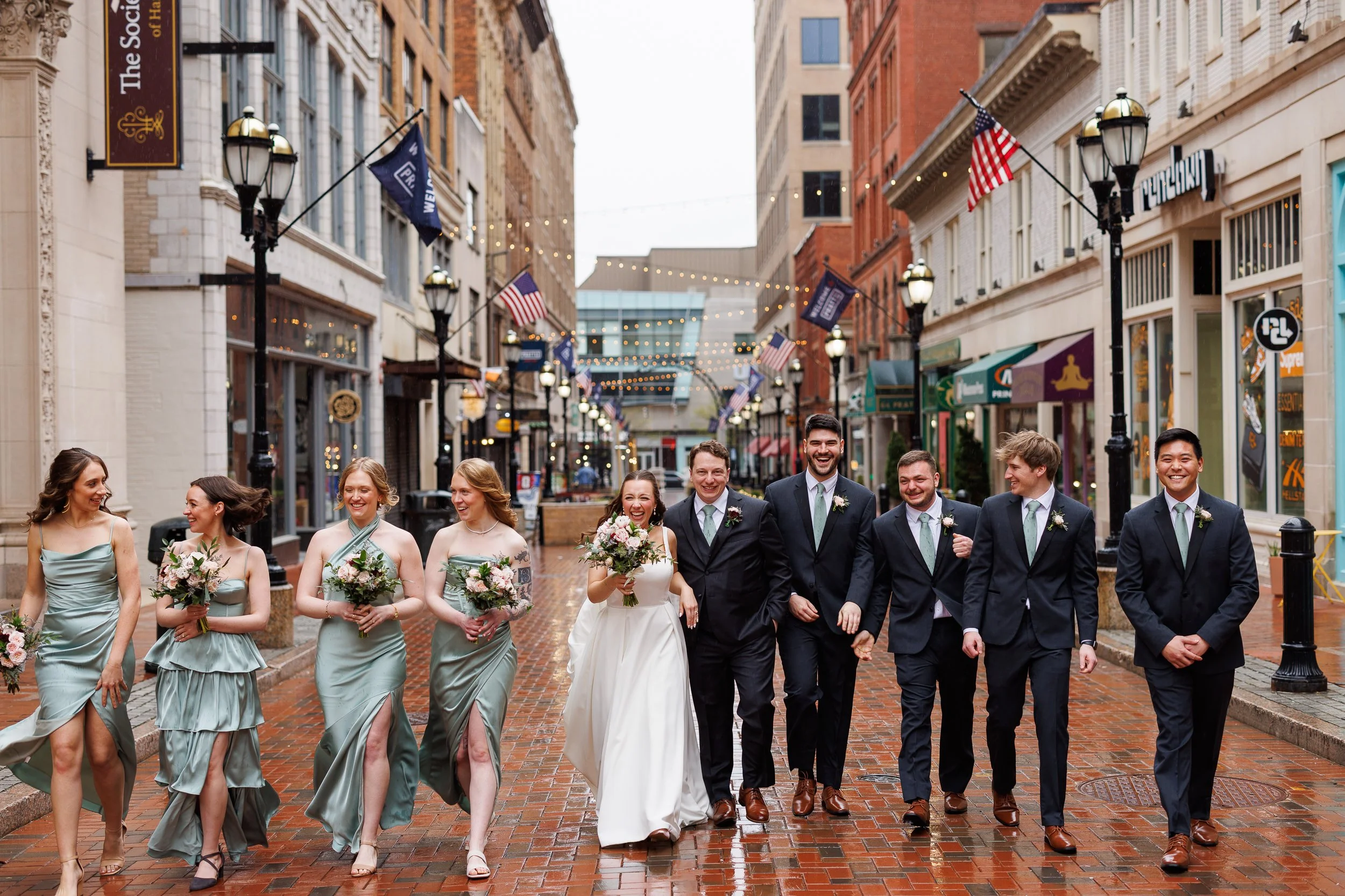 A bride, groom and their wedding party walking down a wet brick street lined with string lights, everyone laughing.