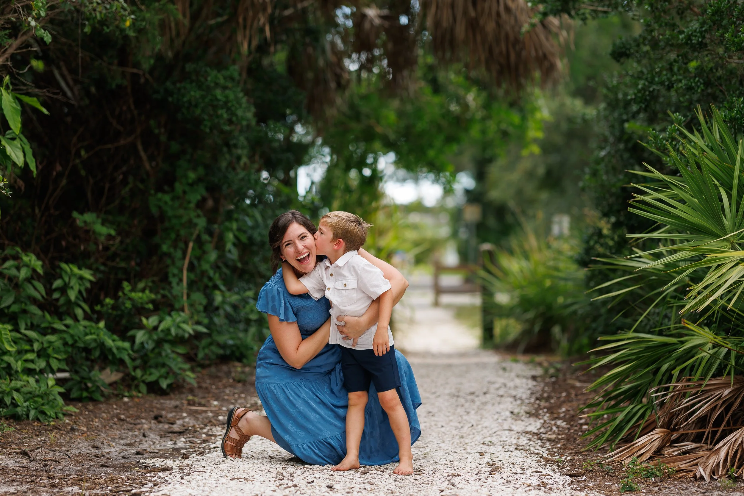 A person in a blue dress kneeling as a child in a white shirt kisses their cheek.