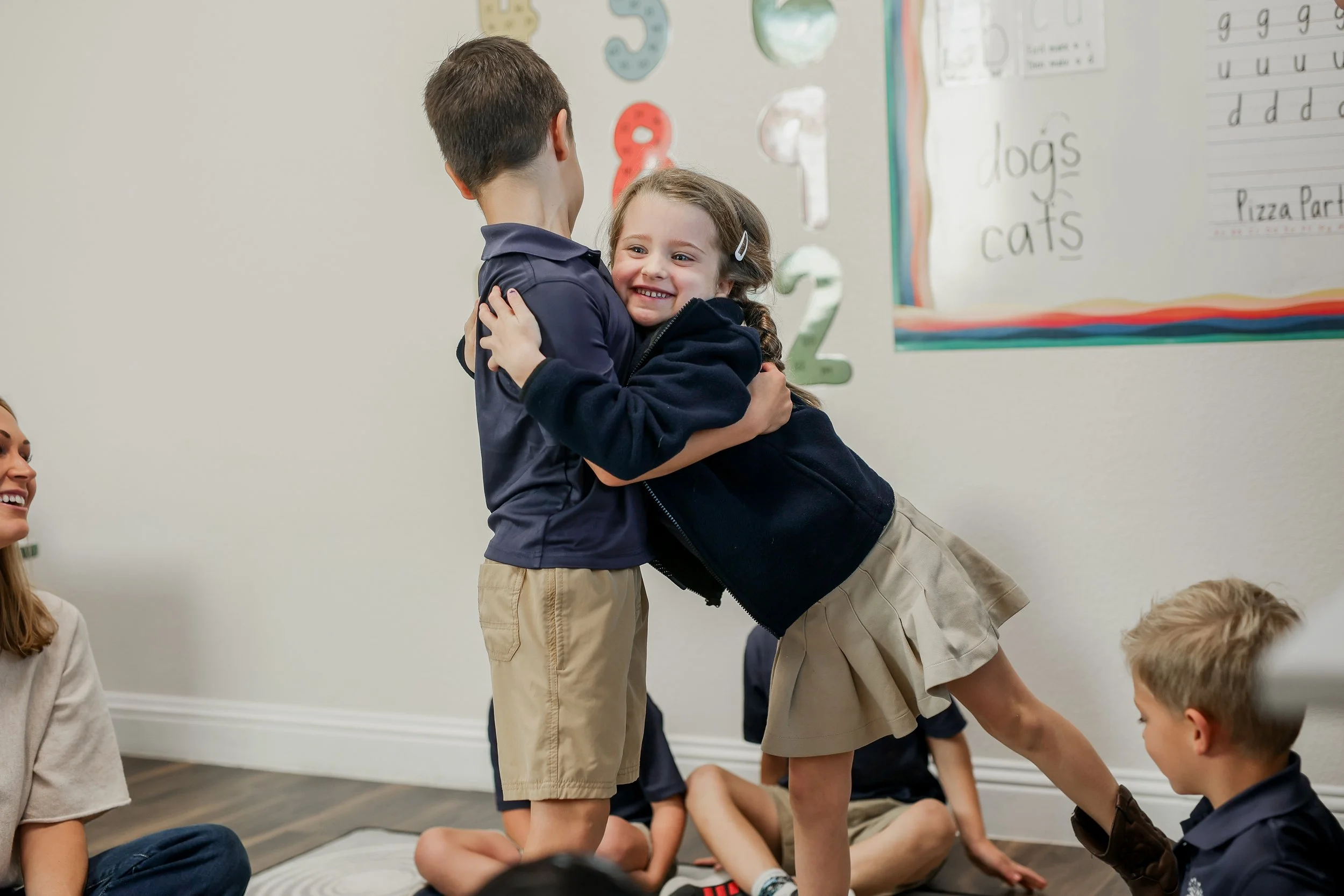 A young girl is hugging a boy in a classroom, both smiling. Other children are seated on the floor around them, and there is a bulletin board with educational materials on the wall.