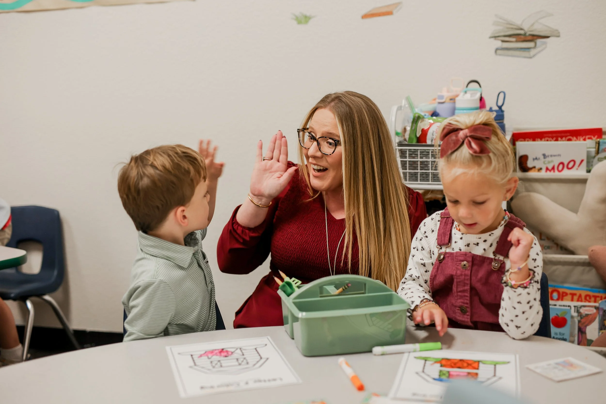 A woman with glasses and long hair giving a high-five to a young boy with brown hair, sitting at a table with coloring sheets. A young girl with a bow in her hair is next to them, looking down. The background shows books, toys, and educational supplies in a classroom or activity room.