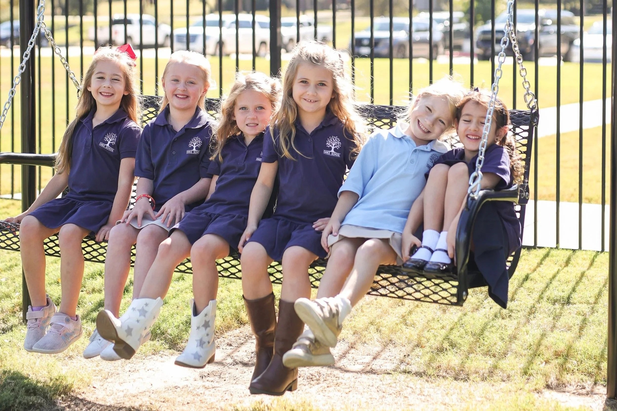 Six young girls sitting together on a black metal swing in a park, smiling, wearing navy blue school uniforms with a tree logo, white and patterned boots, and casual shoes, with a background of green grass, black fence, and parked cars.