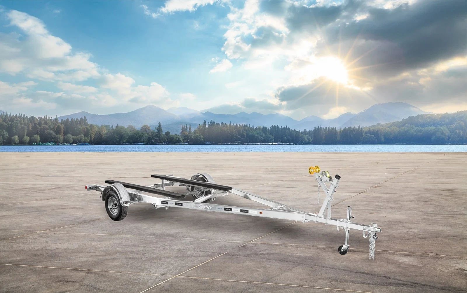 A boat trailer parked on a wide concrete area near a body of water with mountains and trees in the background, under a partly cloudy sky with the sun shining.