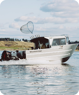 A white motorboat named Wooldridge on calm water with a net attached to a pole on top, under a partly cloudy sky, with land and trees in the background.