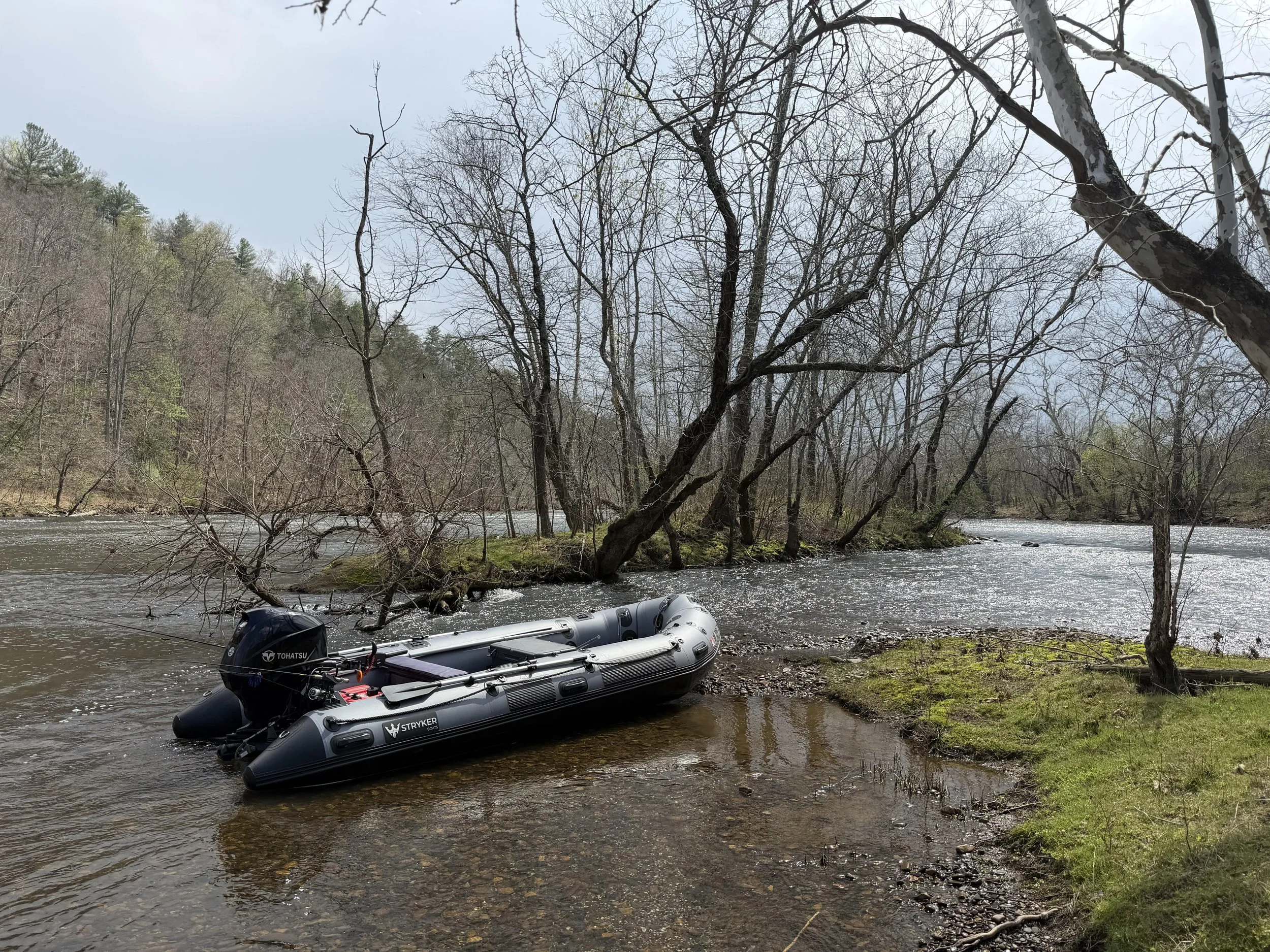 A black inflatable boat with an outboard motor floating near the riverbank of a wooded area with leafless trees and a cloudy sky.