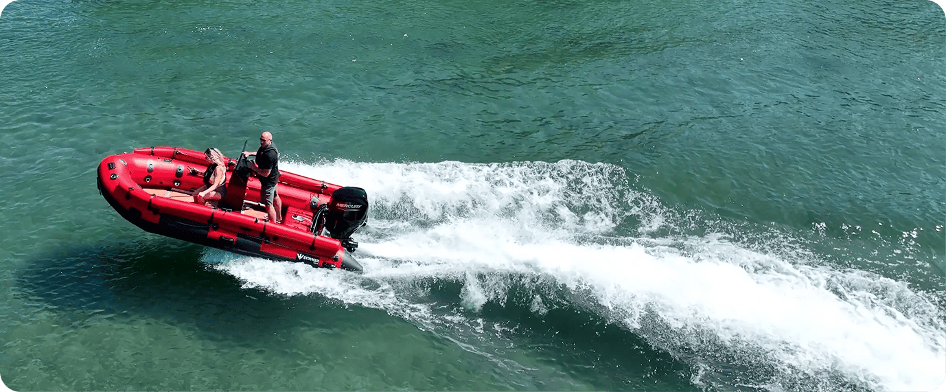 A red inflatable boat speeds across a body of water, creating a wake behind it. Two people are on the boat, one steering and one sitting.