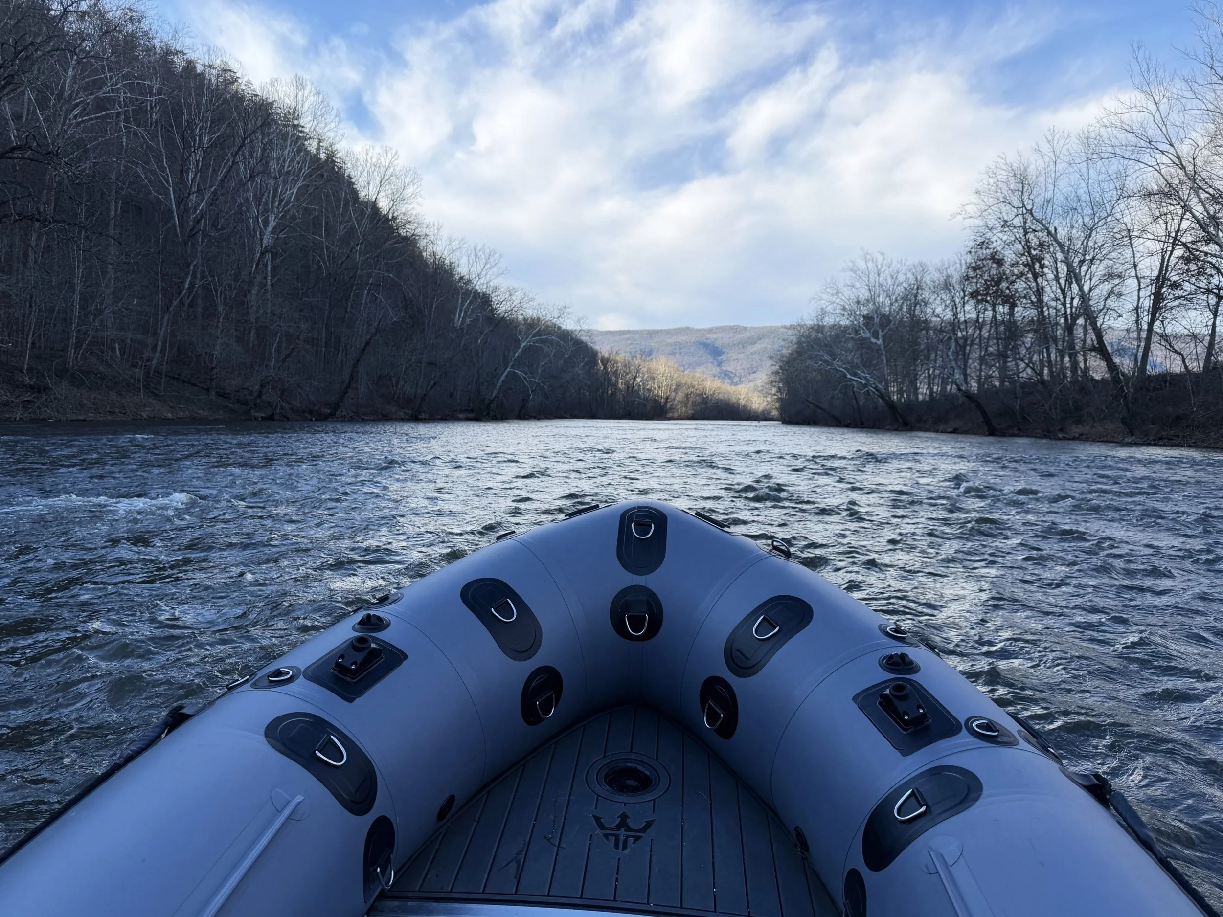 View from a boat on a river, with an inflatable raft in the foreground. The river is surrounded by leafless trees and distant hills under a partly cloudy sky.