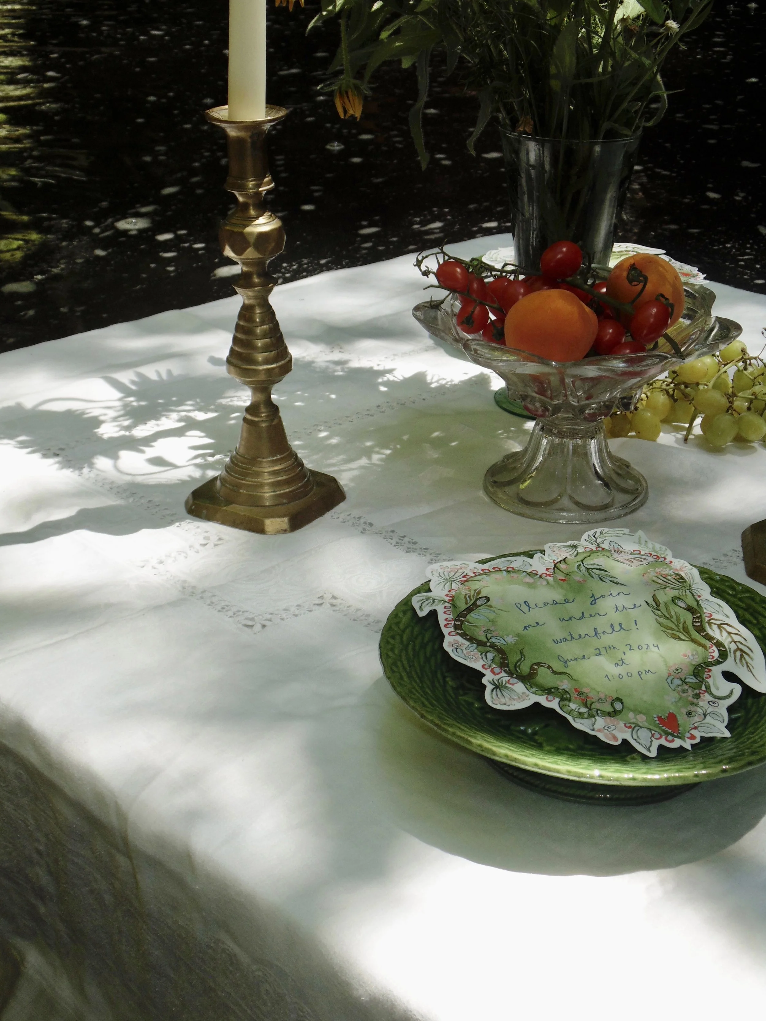 An outdoor table with a white tablecloth, decorated with a gold candlestick, a glass bowl of cherries and tomatoes, a black vase with green foliage, and a green plate with a handwritten note.