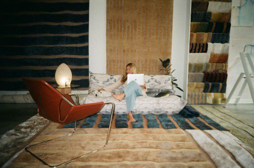 Person sitting on a contemporary sofa, reading a laptop, in a modern, well-decorated living room with various textured rugs, a red armchair, and a bookshelf filled with fabric samples.