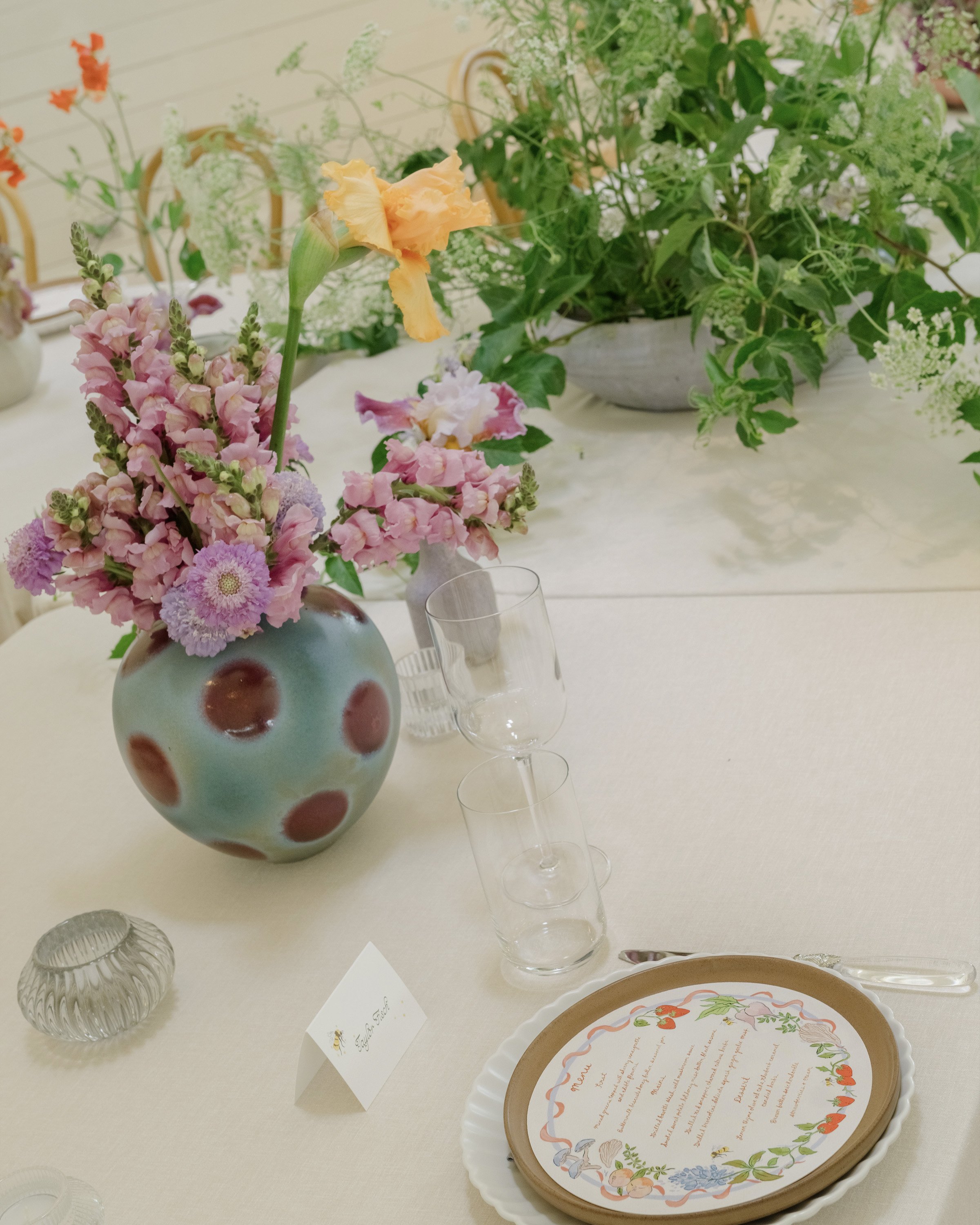 A floral centerpiece on a table with pink, orange, and purple flowers, including a large gladiolus, in a polka-dotted vase, with an empty wine glass and a decorative plate with handwritten menu text, in a well-lit room decorated with green foliage.