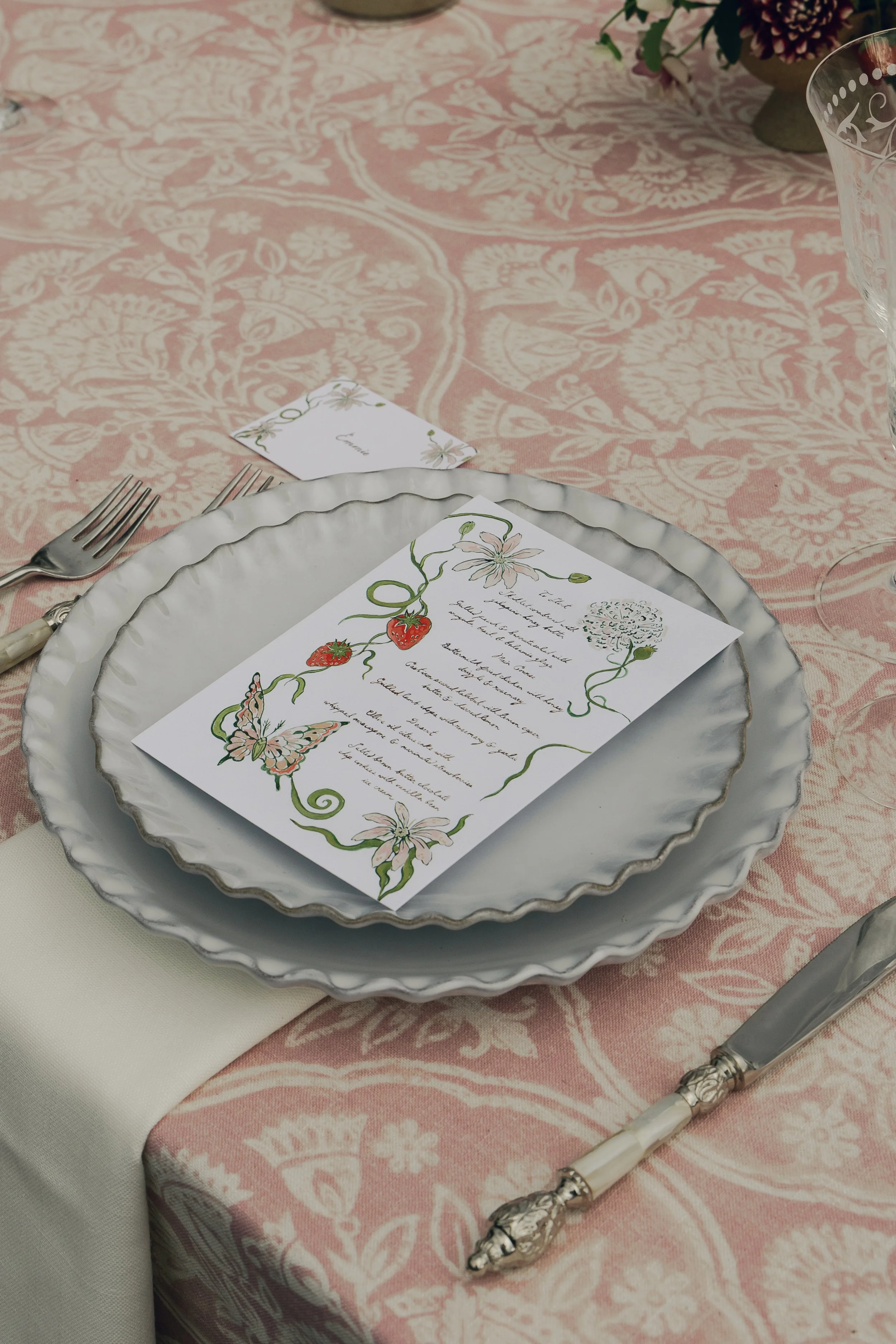 Elegant table setting with a pink and white patterned tablecloth, a white dinner plate with a decorative scalloped edge, a handwritten menu card with floral and butterfly illustrations, a matching place card, and silverware with ornate handles.