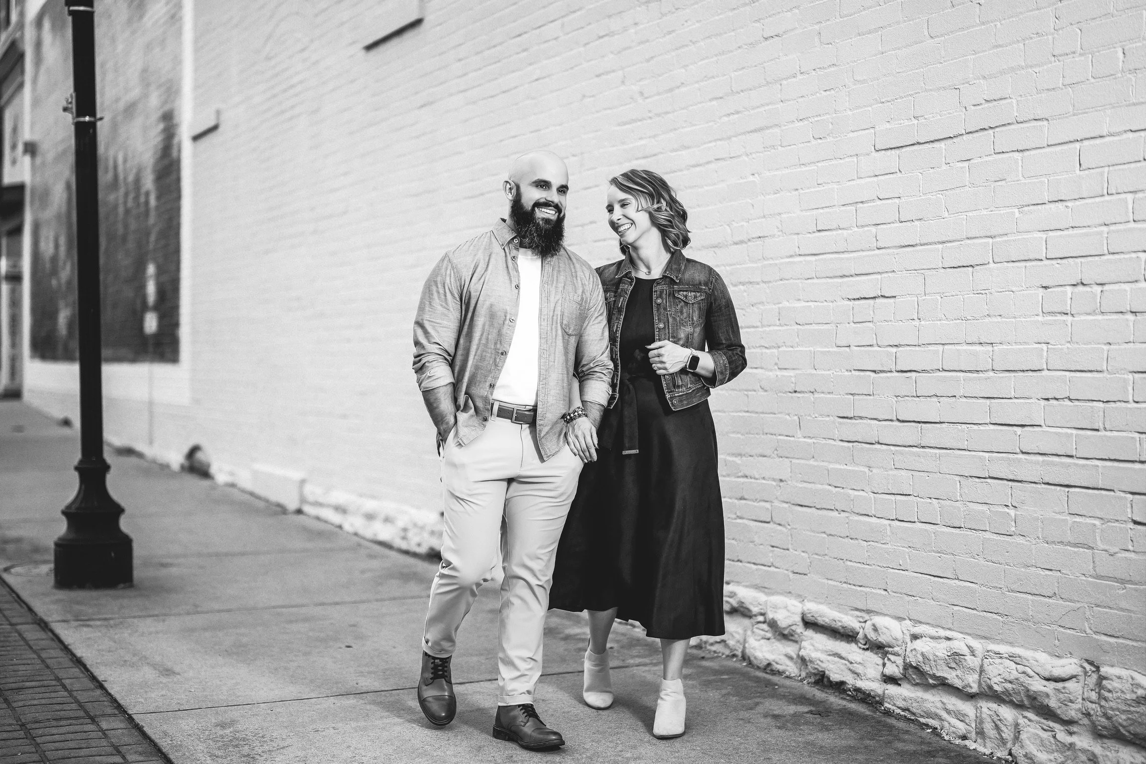 A man and woman walking on a city sidewalk, smiling and talking, with a brick wall in the background.