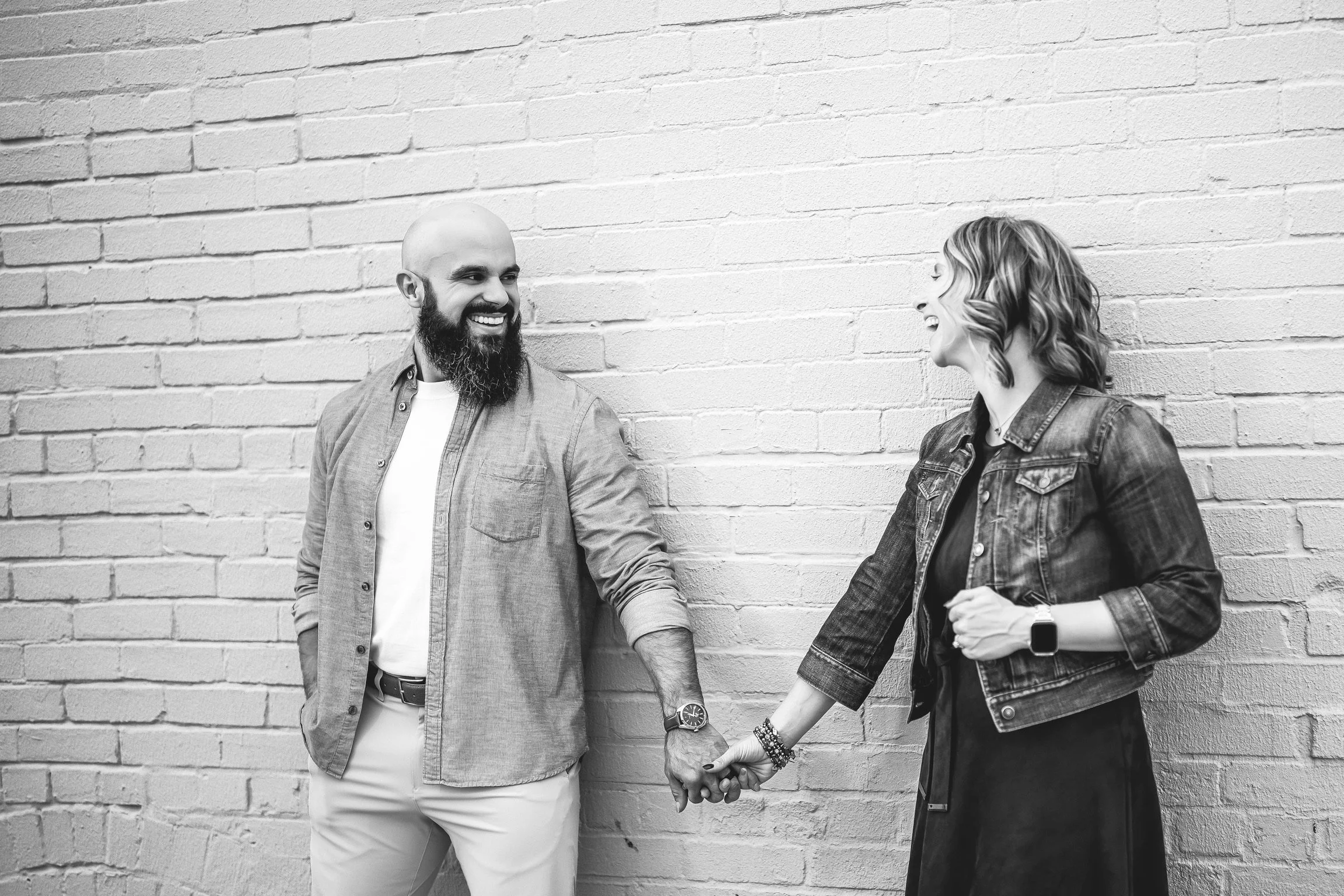 A black-and-white photo of a man and woman holding hands and laughing against a brick wall. The man has a beard, is bald, and wears a button-up shirt over a T-shirt. The woman has shoulder-length hair, wears a denim jacket and a dress, and has a smartwatch on her wrist.