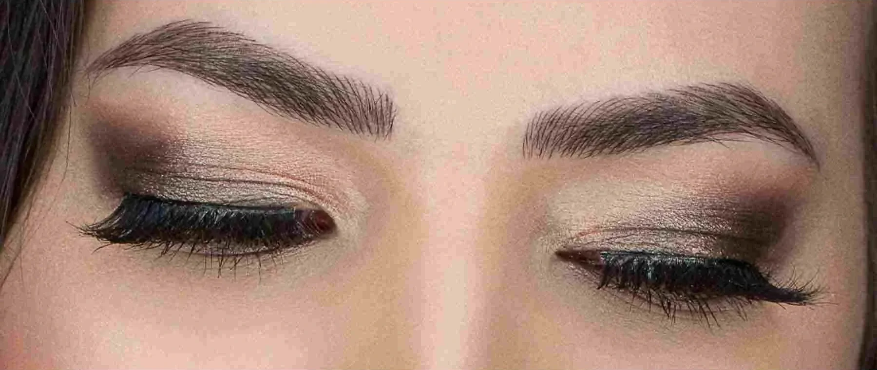 Close-up of a woman's closed eyes showcasing well-defined eyebrows, eyeshadow in neutral tones with shimmer, black eyeliner, and long mascara-coated eyelashes.