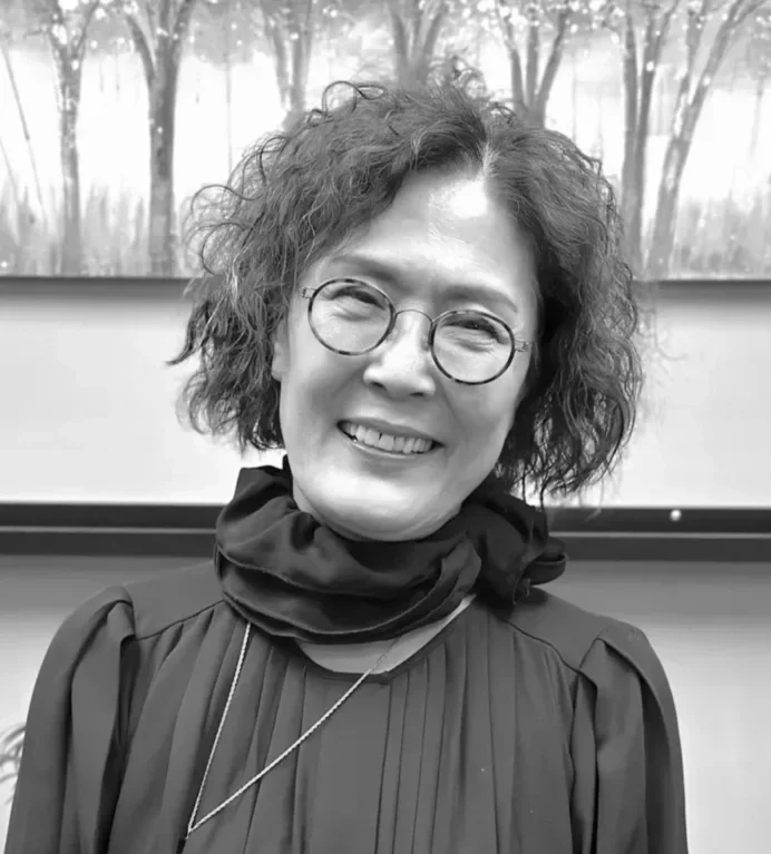 Hair stylist, Moon. A woman with short curly hair, round glasses, and a scarf, smiling in front of a framed picture of trees.