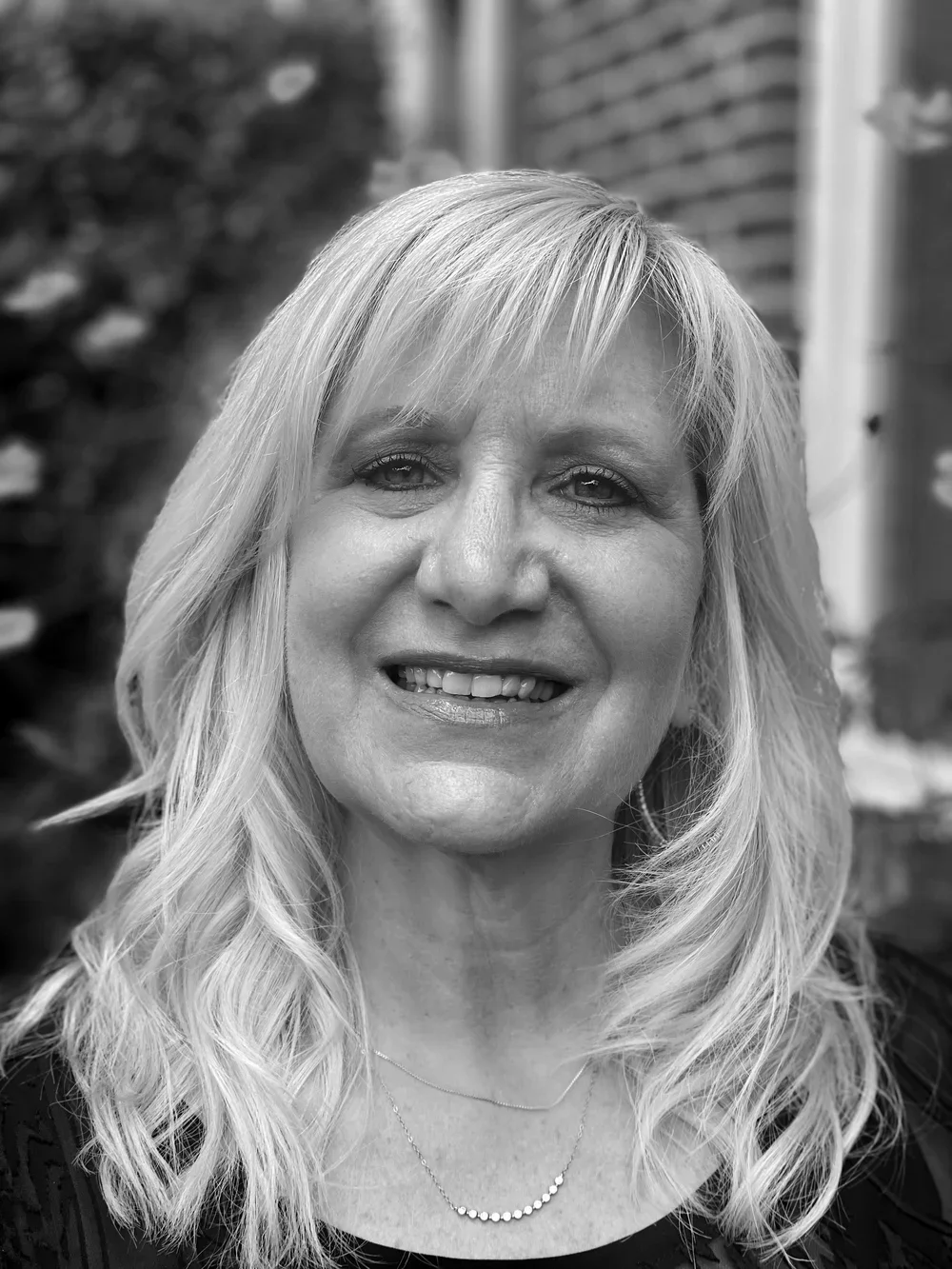 Hair stylist, Darlene. A woman with wavy, shoulder-length blonde hair, smiling in front of a brick wall and some foliage.