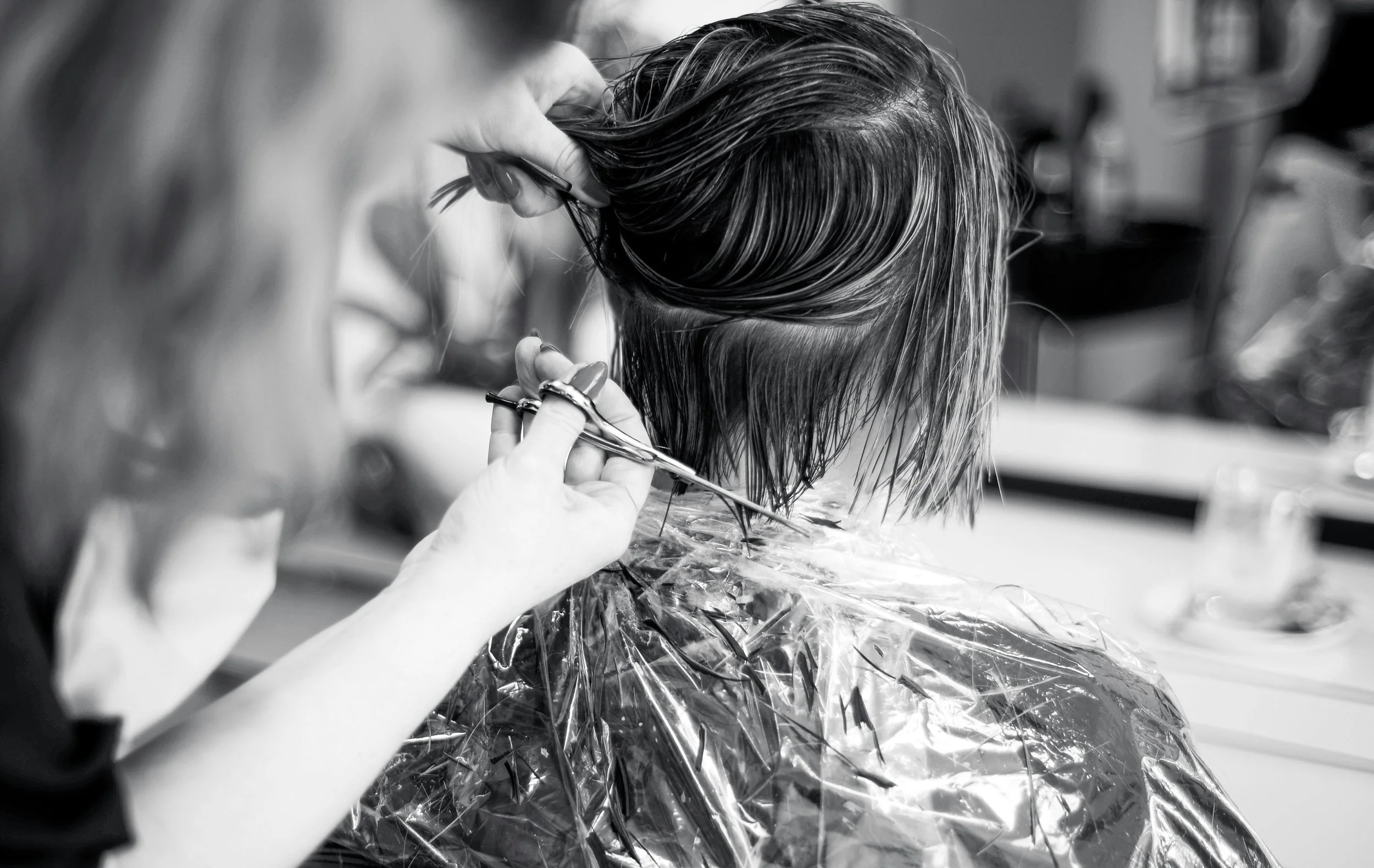 A stylist cuts a person's wet hair with scissors in a salon. The person is covered with a plastic cape.