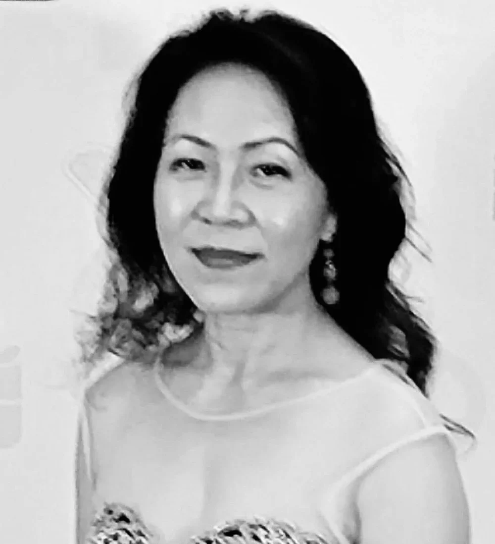 Beauty professional, Sung. A woman with dark wavy hair, earrings, wearing a sleeveless top with a patterned design, smiling slightly.