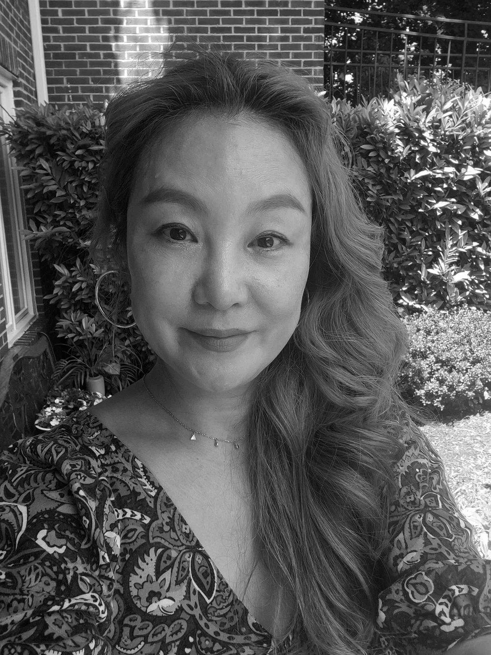 Hair stylist, Cecilia. A woman with long wavy hair, wearing hoop earrings and a patterned top, smiling outdoors in front of bushes and a brick building.