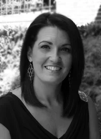 Hair stylist, Amy. A woman with shoulder-length hair, smiling, wearing earrings and a dark top, outdoors in front of trees.