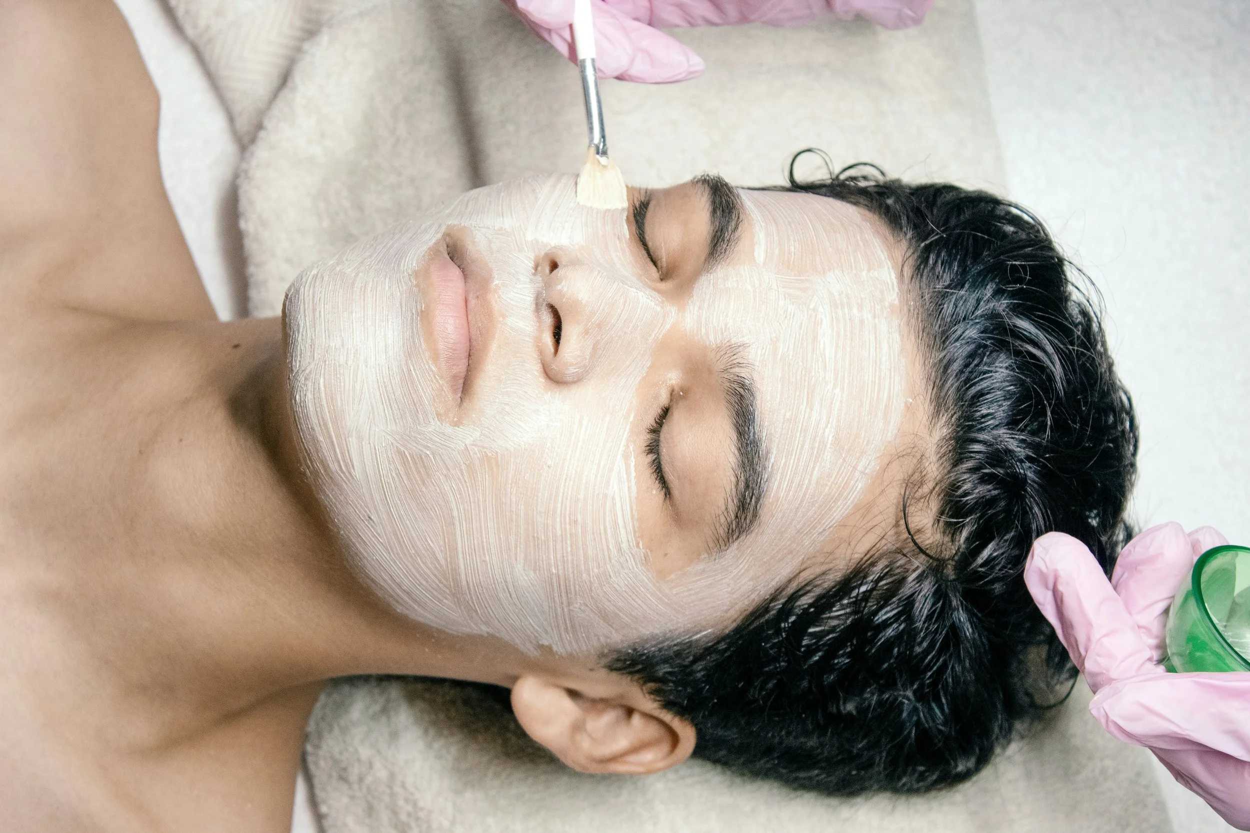 A person receiving a facial treatment with a white mask applied on their face, lying down with eyes closed, while a skincare professional applies a substance with a small brush.