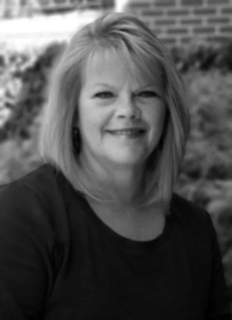 Hair stylist, Arlene. A woman with shoulder-length blonde hair smiling outdoors, with a brick wall and shrubbery in the background.
