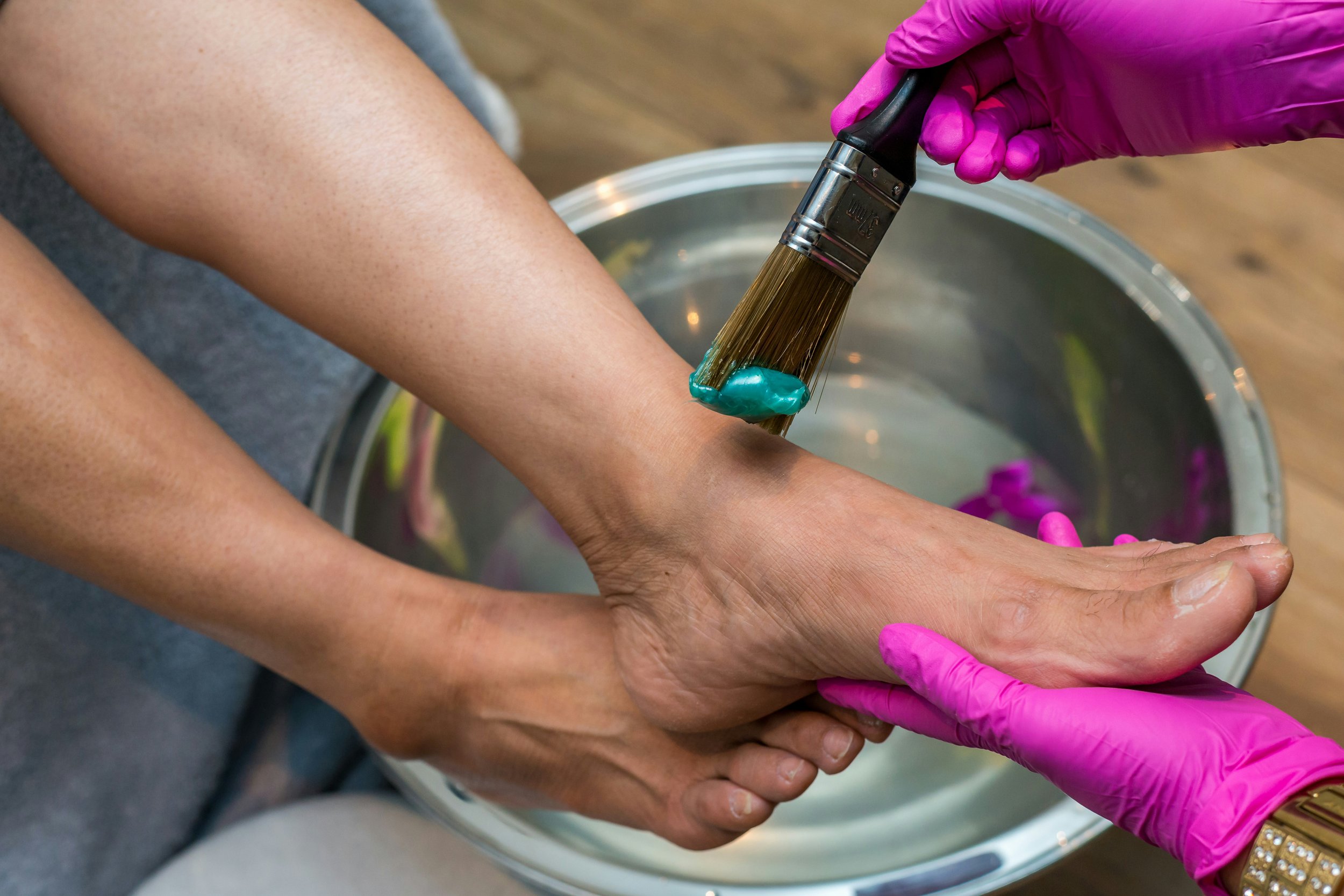 Person receiving a pedicure, with a shiny teal nail polish being applied to their toenail using a brush, while wearing pink gloves, over a bucket of water.