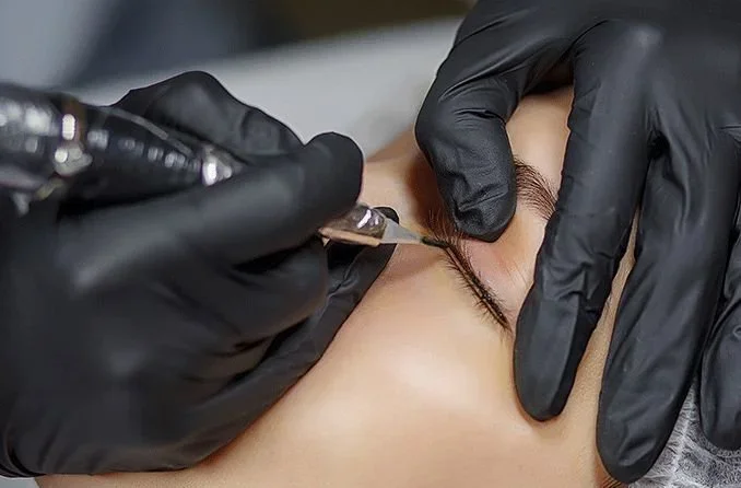 A person receiving an eyeliner tattoo procedure from a technician wearing black gloves, with a focus on the eye and technician's hands