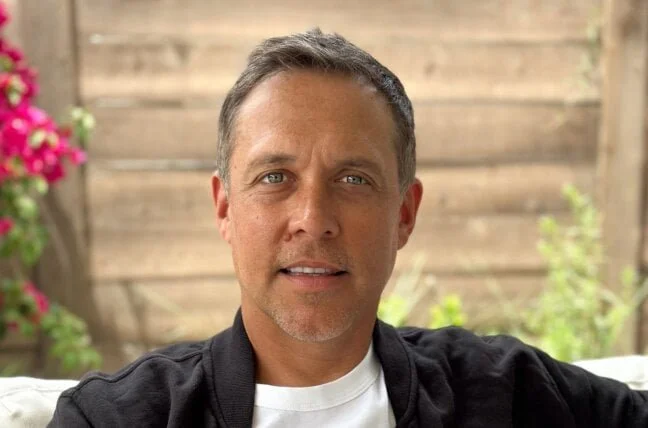 A man with short, dark hair and light skin taking a selfie outdoors, with a wooden fence and pink flowers in the background.