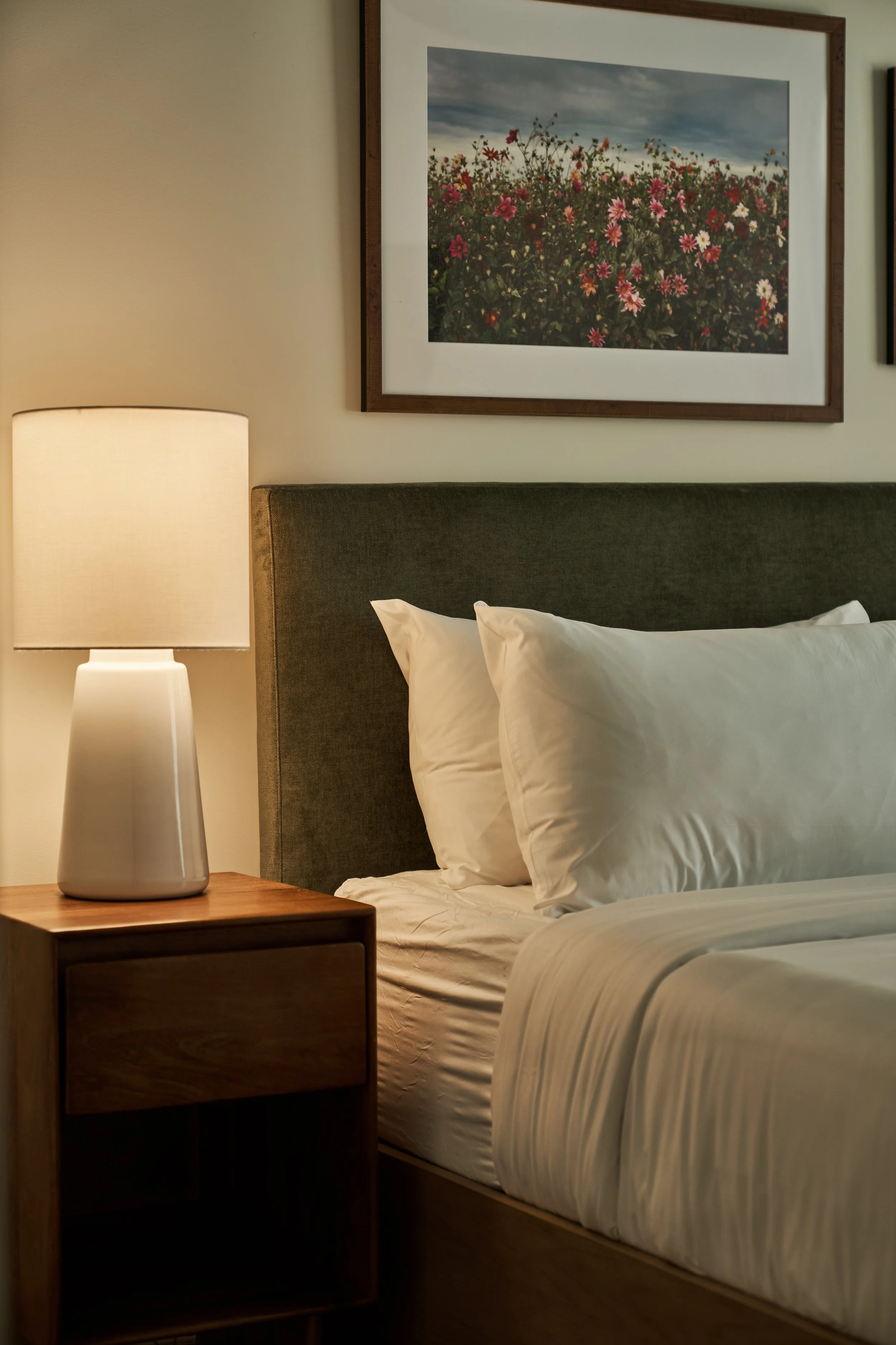 A bedroom scene with a bed, white pillows, a wooden nightstand, a table lamp, and a framed picture of flowers on the wall.
